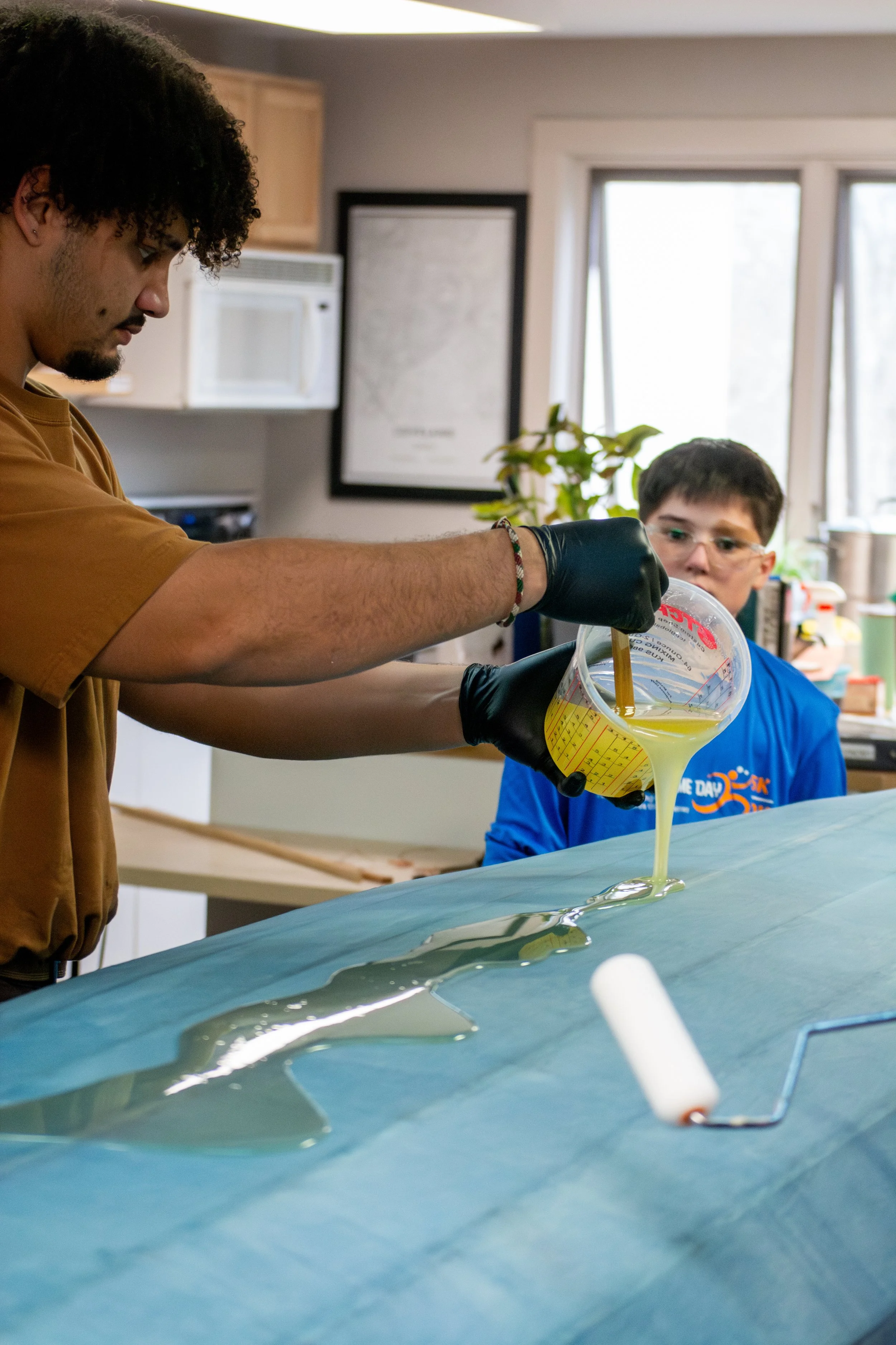 A person pouring a yellow liquid from a measuring cup onto a large blue sheet, with a young boy observing in the background.