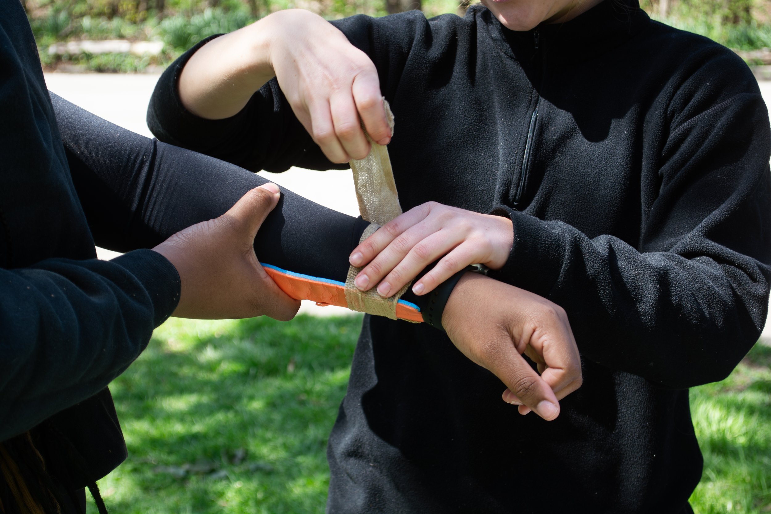 Two people assisting each other with first aid in an outdoor setting, one person applying a bandage to the other's arm.