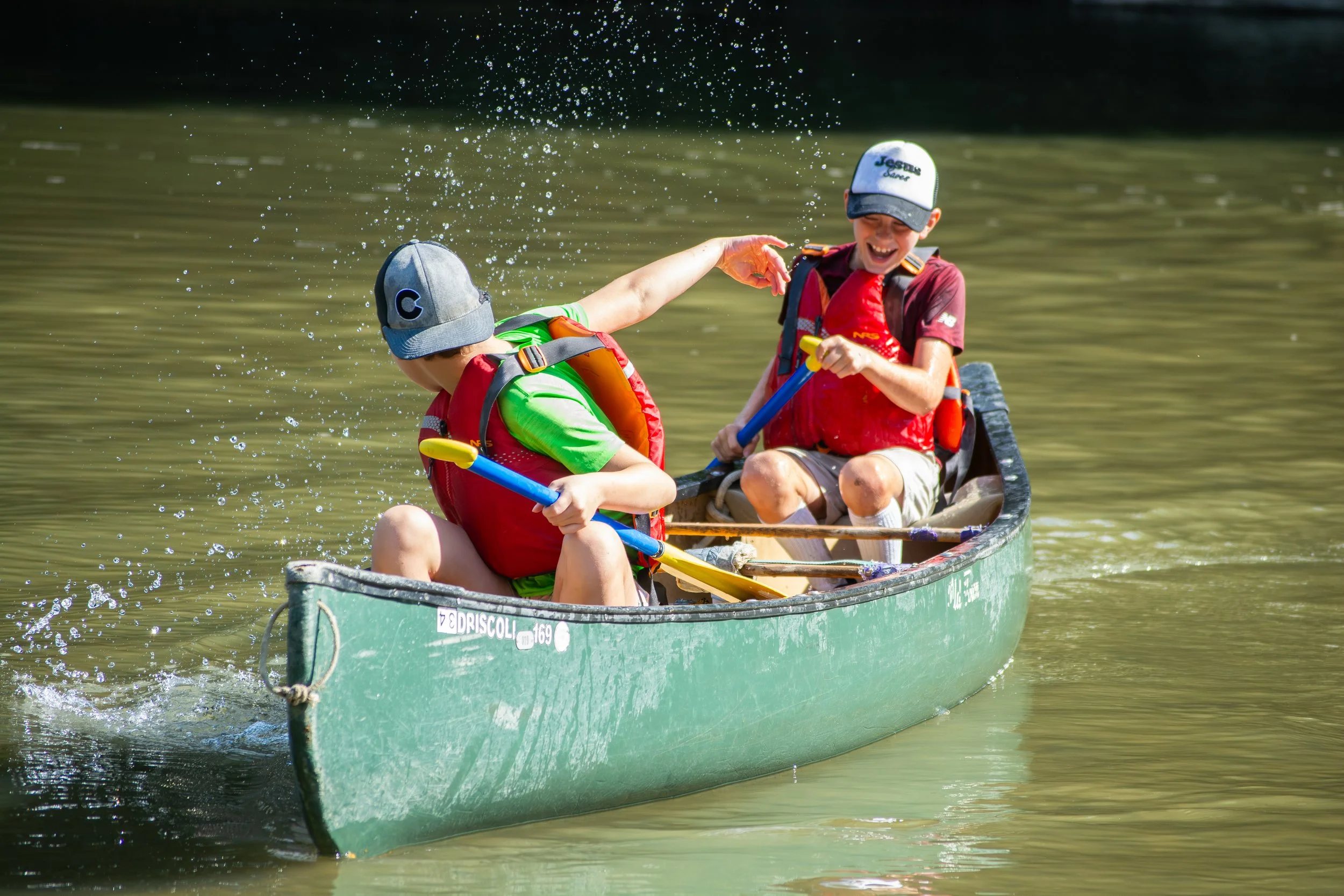 Two boys playfully splashing water at each other while sitting in a small green boat on a lake, wearing life jackets and hats.