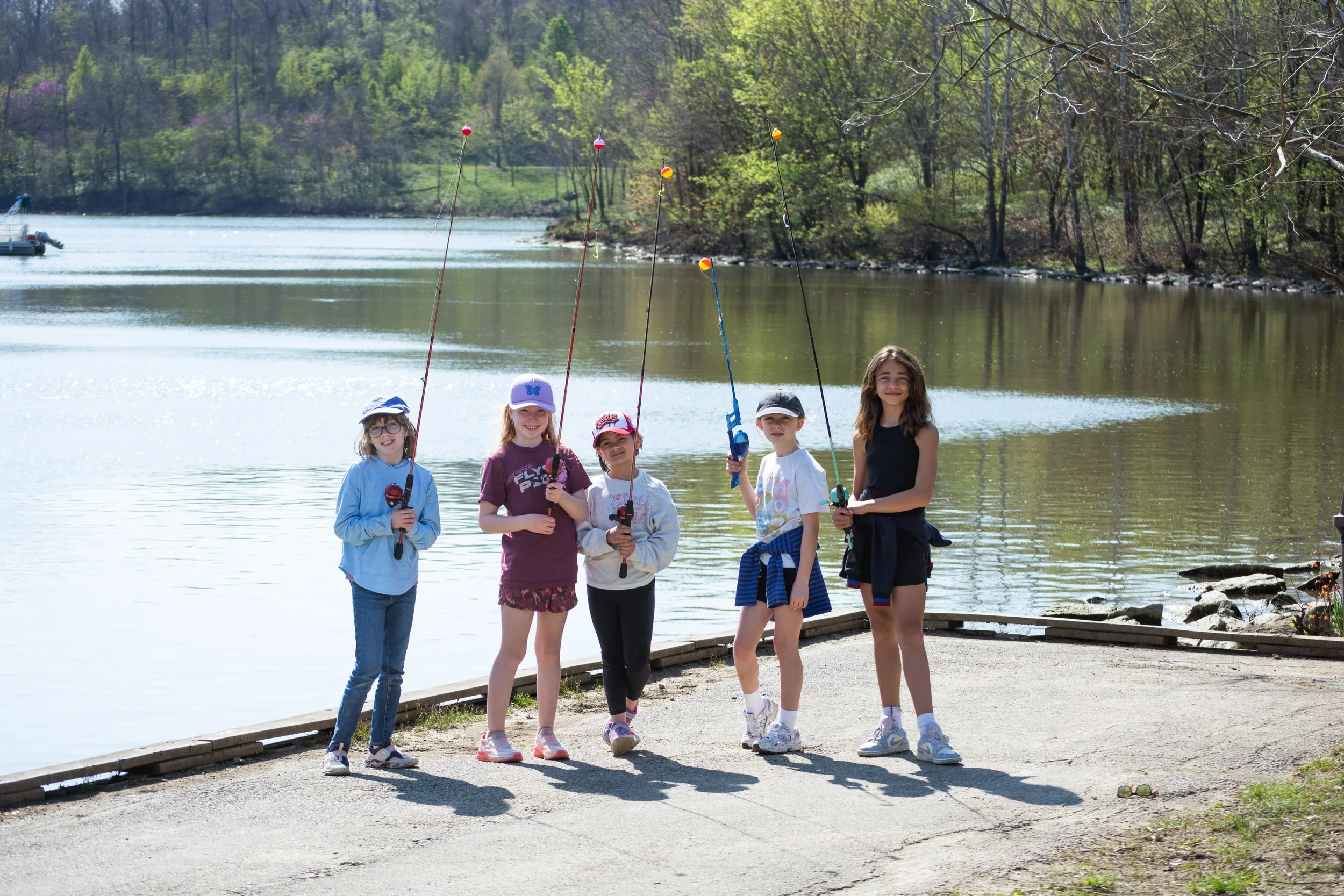 Five children, four girls and one boy, standing on a dock by a lake holding fishing poles, smiling, with trees and a boat in the background on a sunny day.