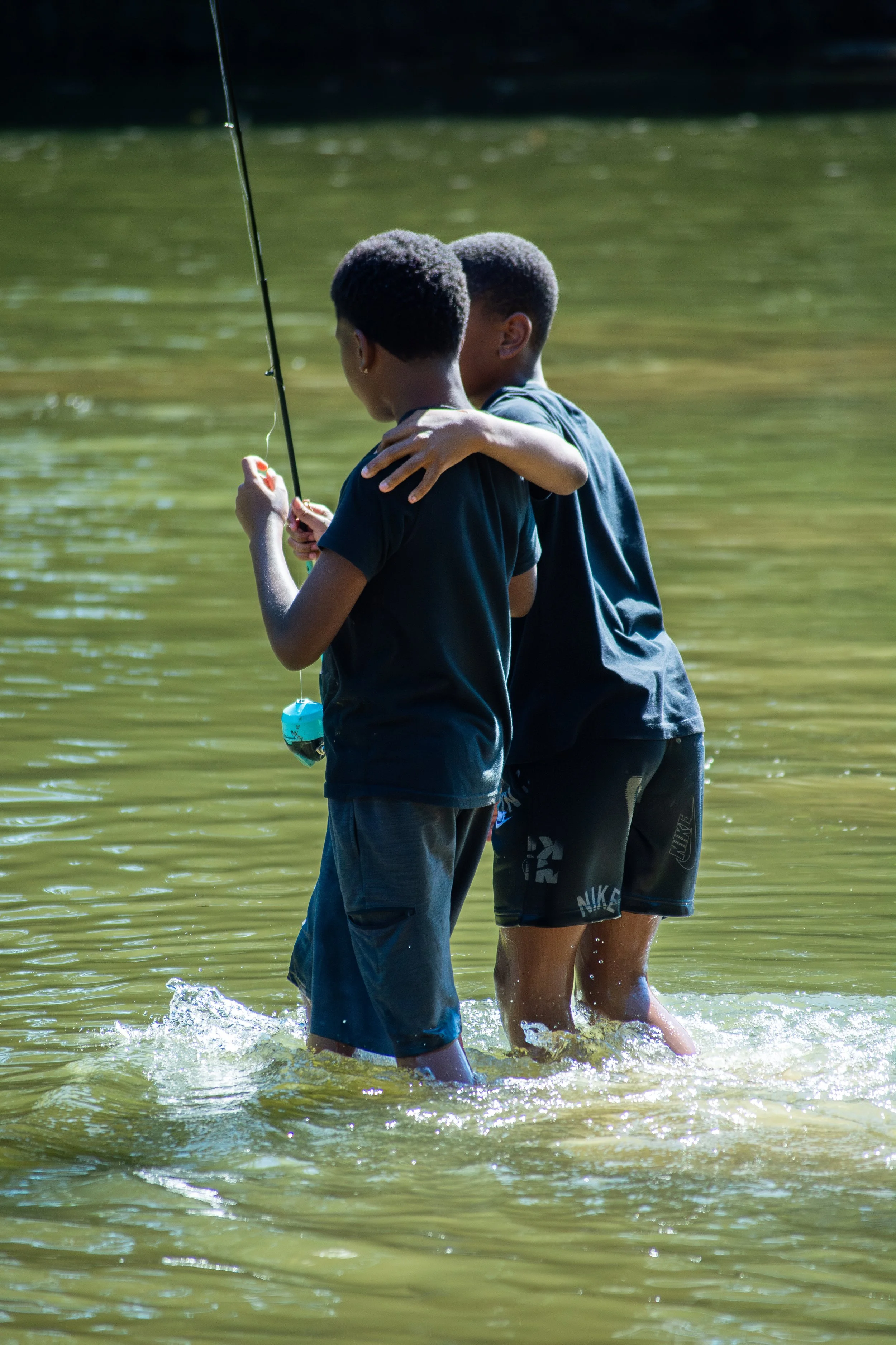 Two boys standing in water, embracing each other, next to a person holding a fishing rod.