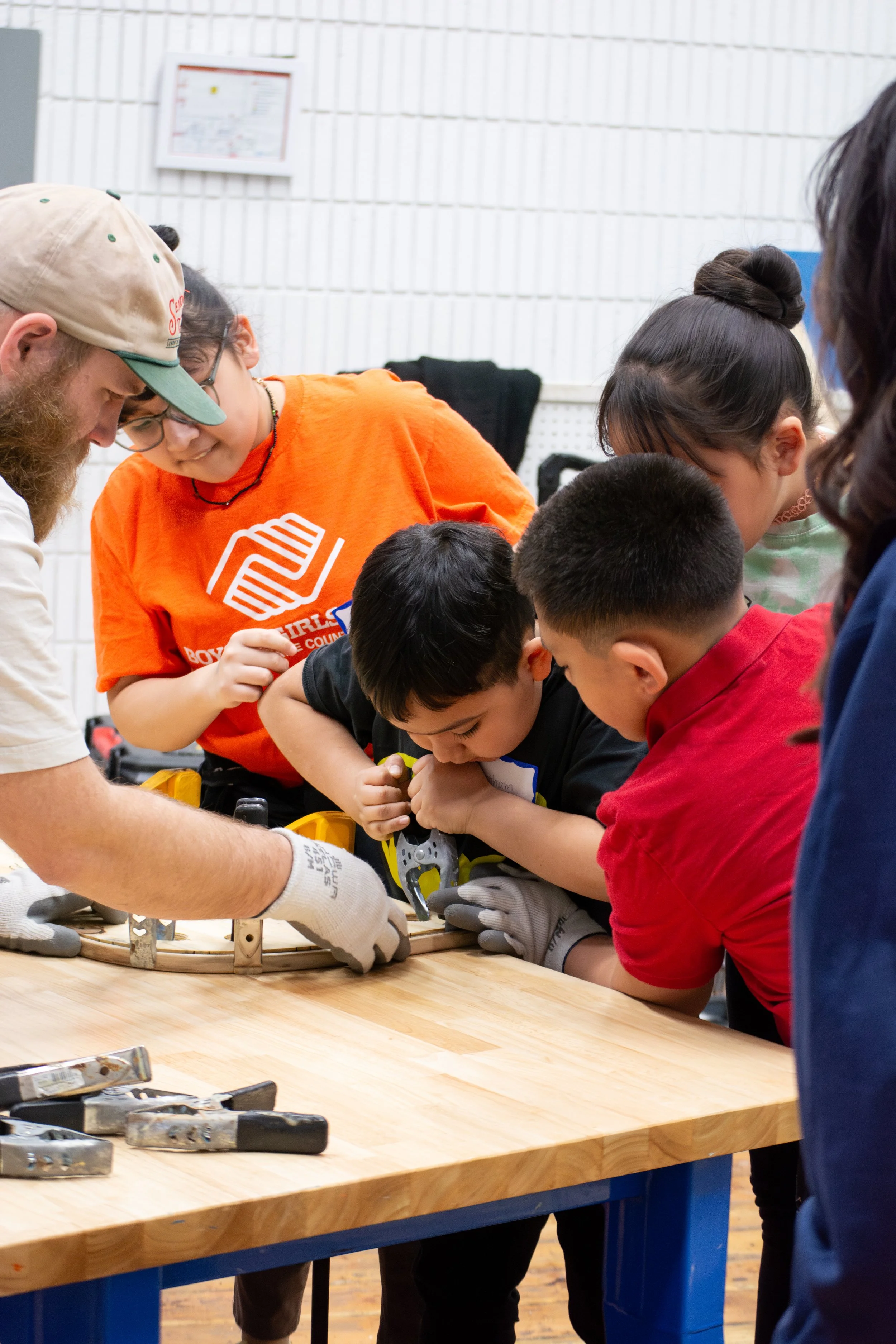 A group of children and adults working together on a woodworking project in a workshop. They are gathered around a wooden workbench, using tools to work on a circular wooden piece. Several tools are laid out on the table.