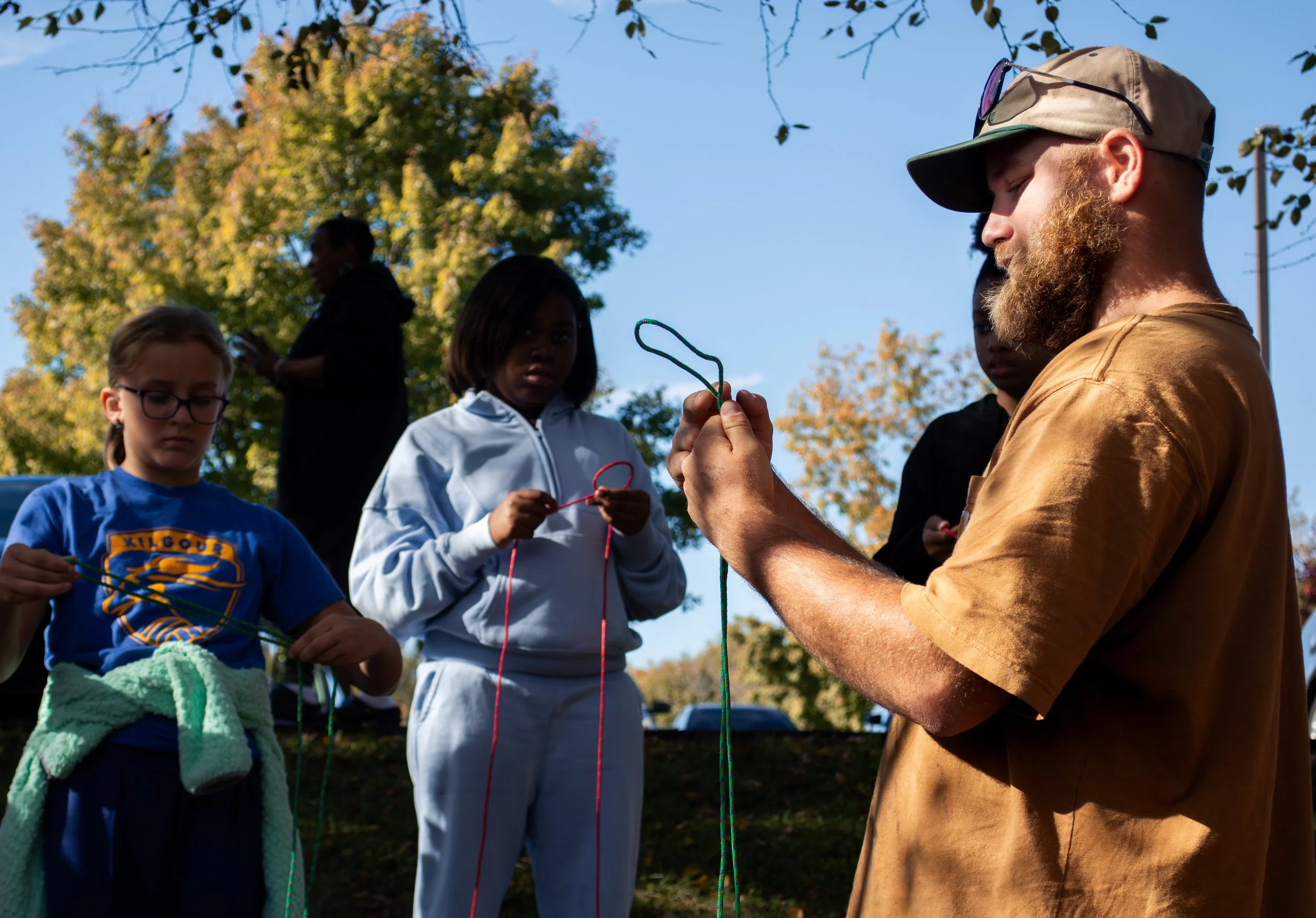 A group of children and an adult outdoors under trees, preparing with colorful ropes or cords, possibly for an outdoor activity or team-building exercise, with a clear blue sky in the background.