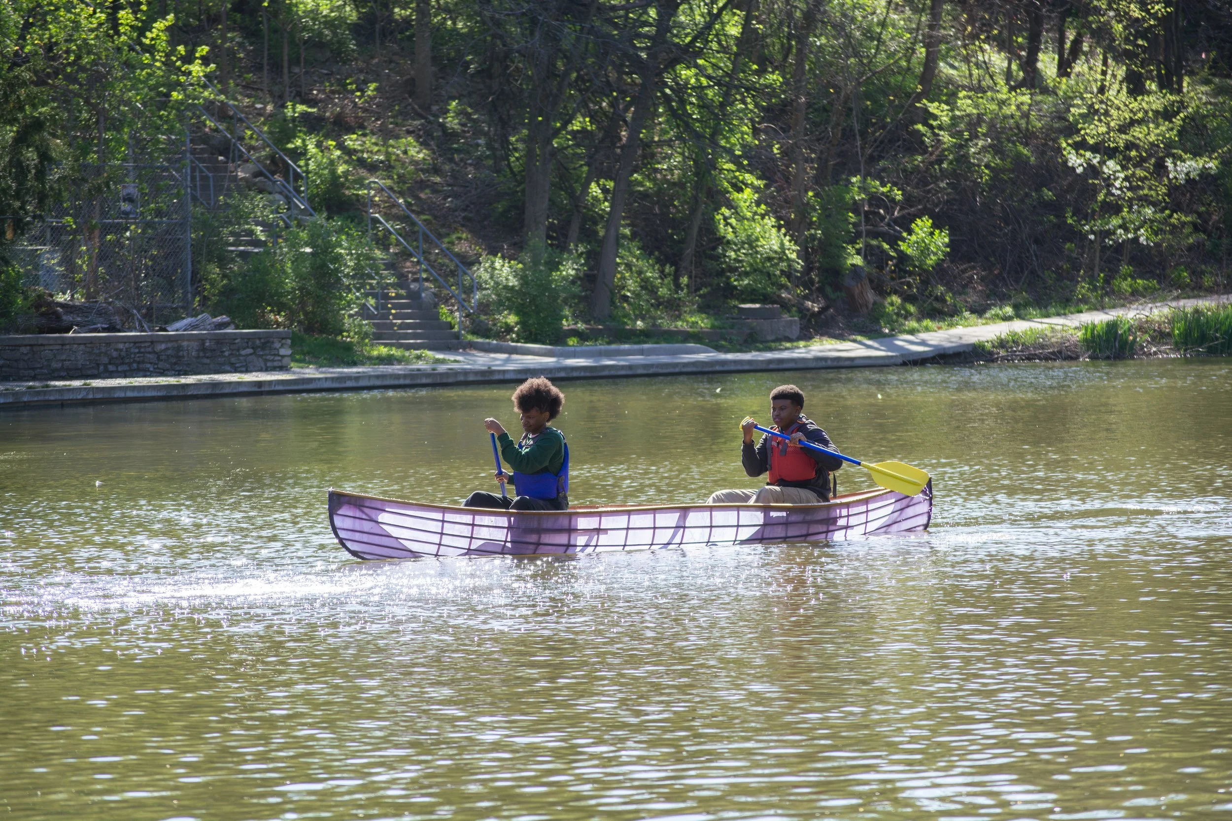 Two children kayaking on a calm river with lush green trees and a stone staircase in the background.