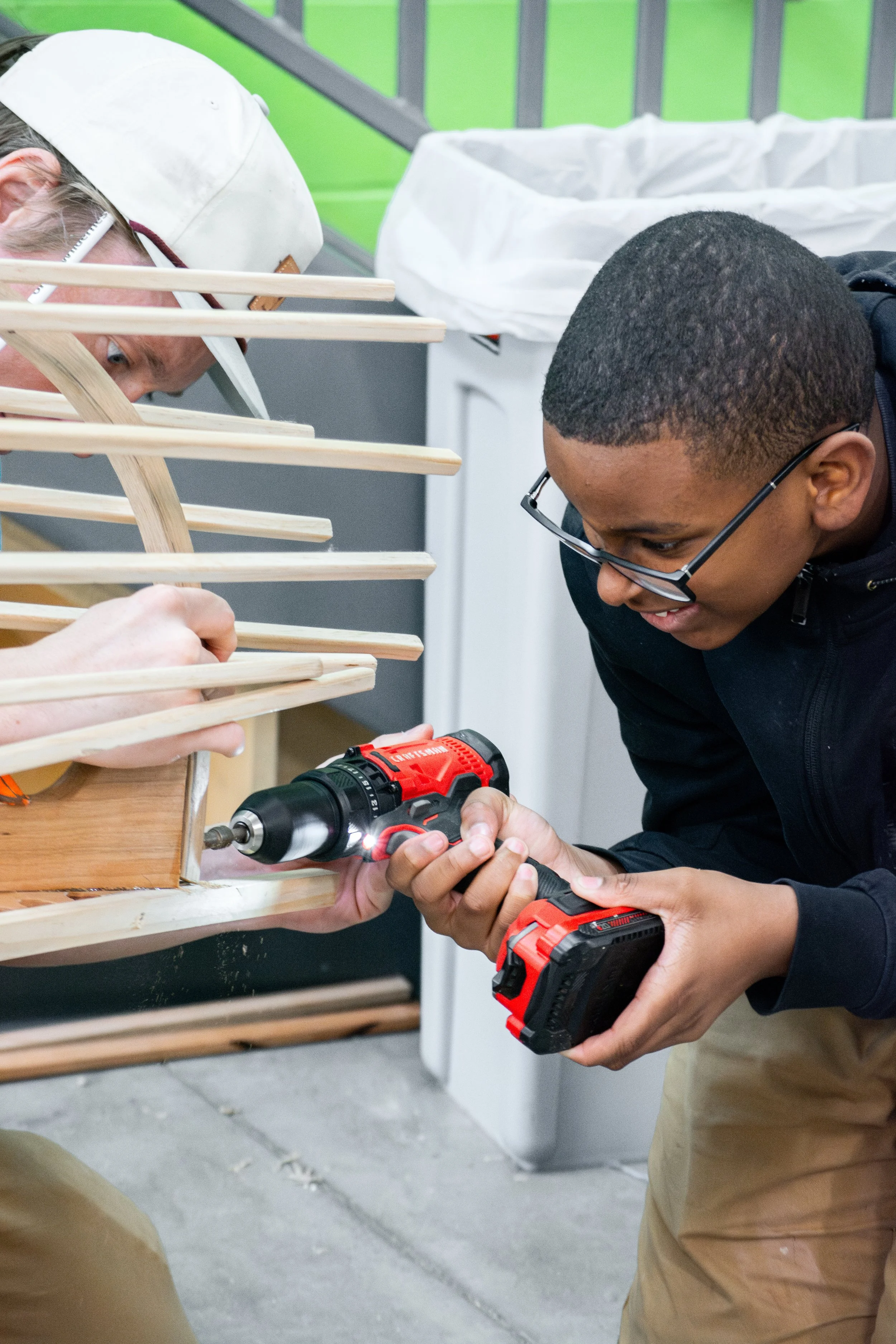 Two men working together on a woodworking project in a workshop, one holding a cordless drill and the other guiding it, surrounded by wooden pieces and tools.