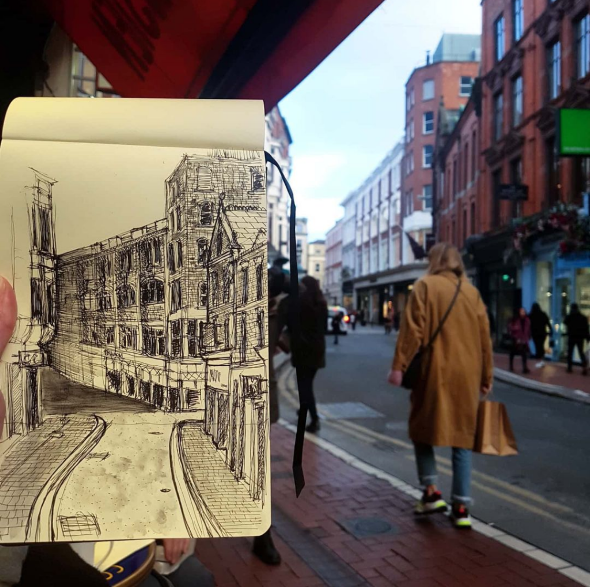 A hand holding a sketchbook with a drawing of a city street scene, with buildings and a curve in the road, in front of a real street scene with pedestrians and multi-story buildings.
