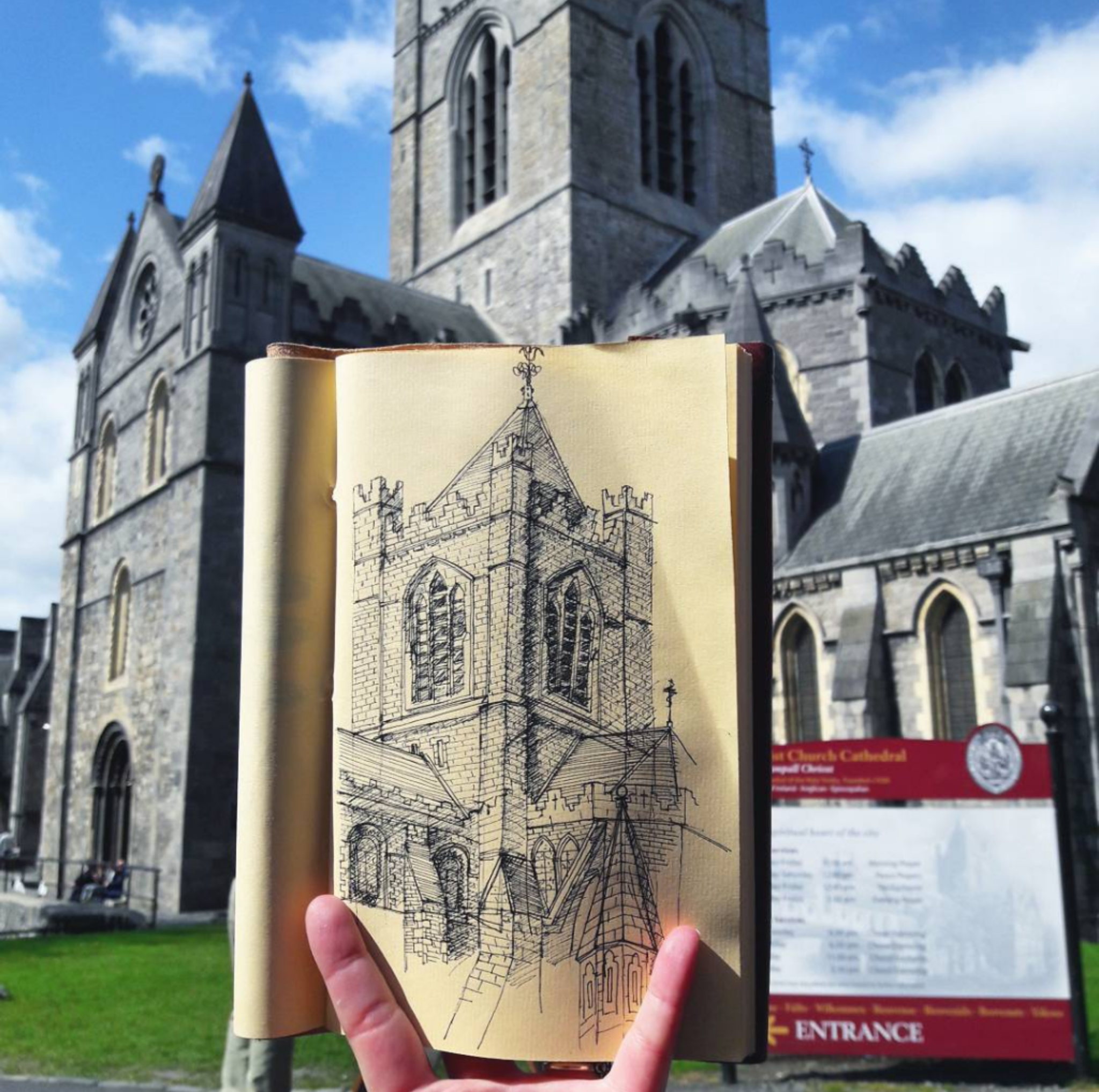 A hand holding an open sketchbook with a drawing of a church tower, in front of a real church building under a partly cloudy sky.