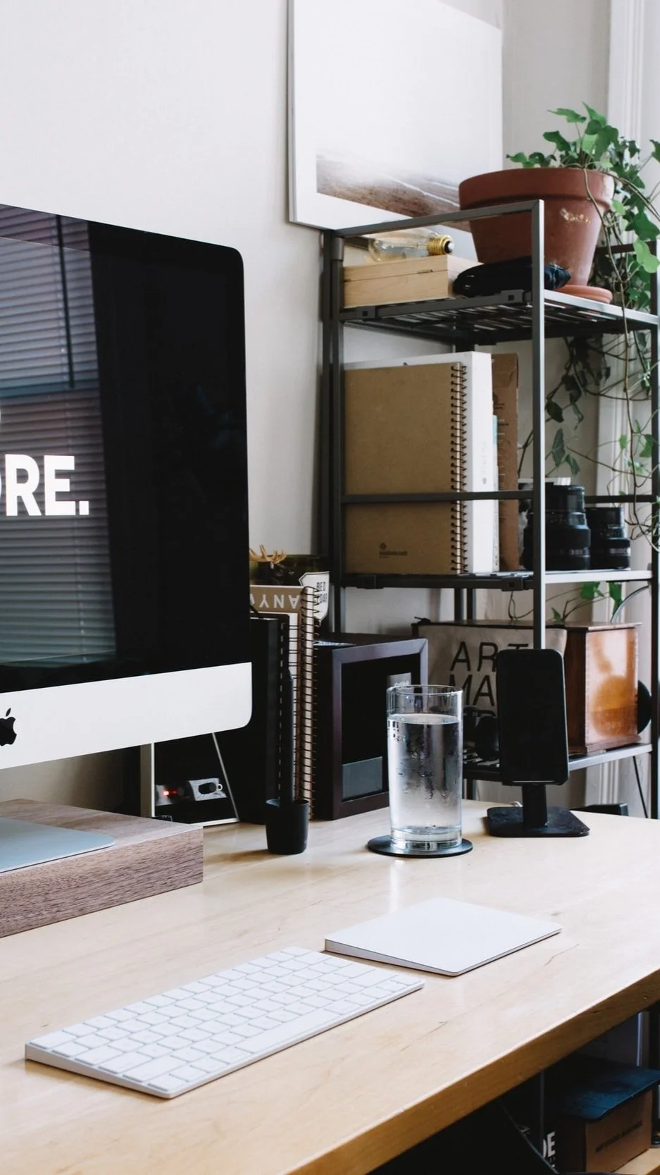 Modern workspace with desktop computer, keyboard, glass of water, and office shelving with books, plants, and decor.