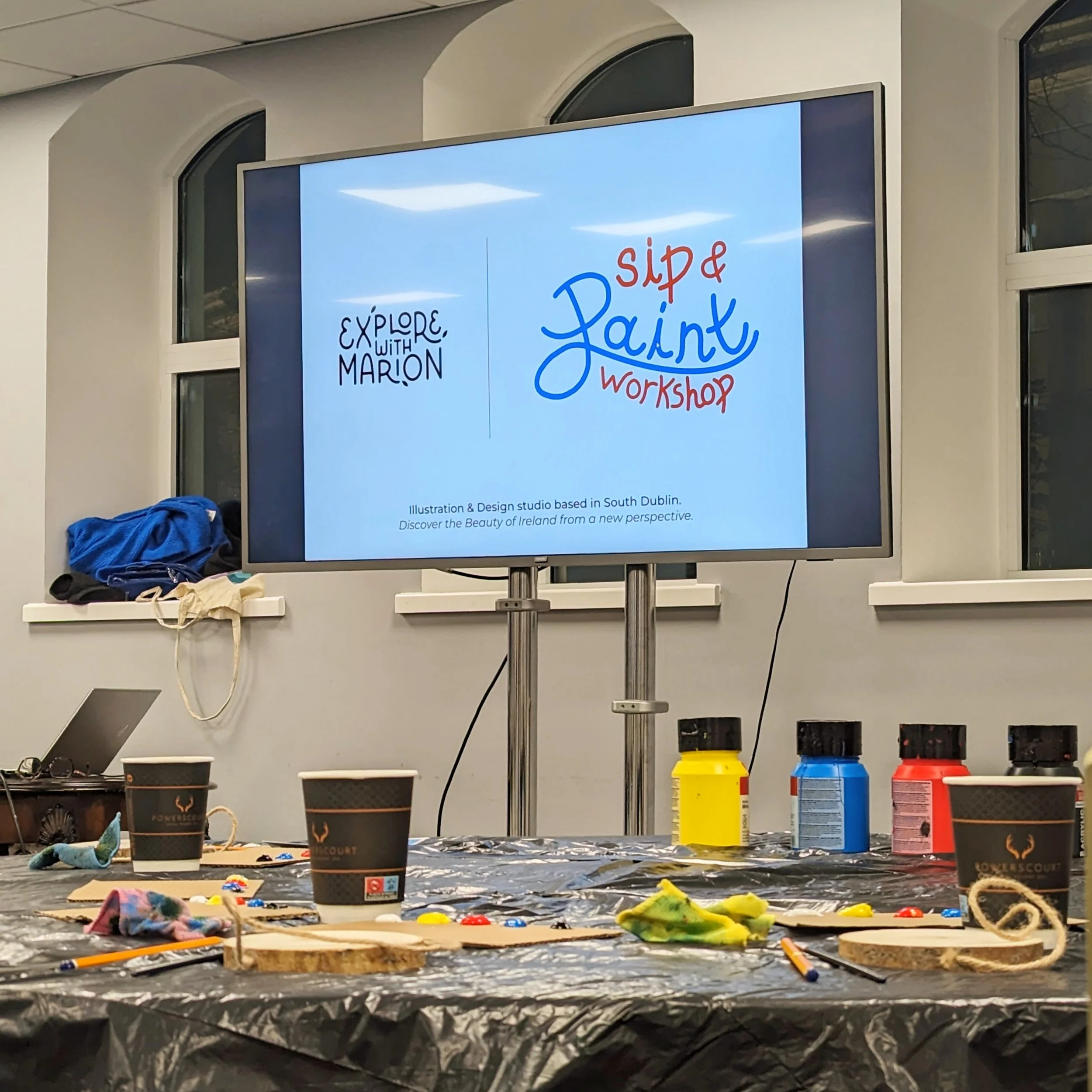 Sip and Paint workshop setup with a screen displaying workshop information, art supplies like paints, brushes, and cups on a table covered with a black cloth.