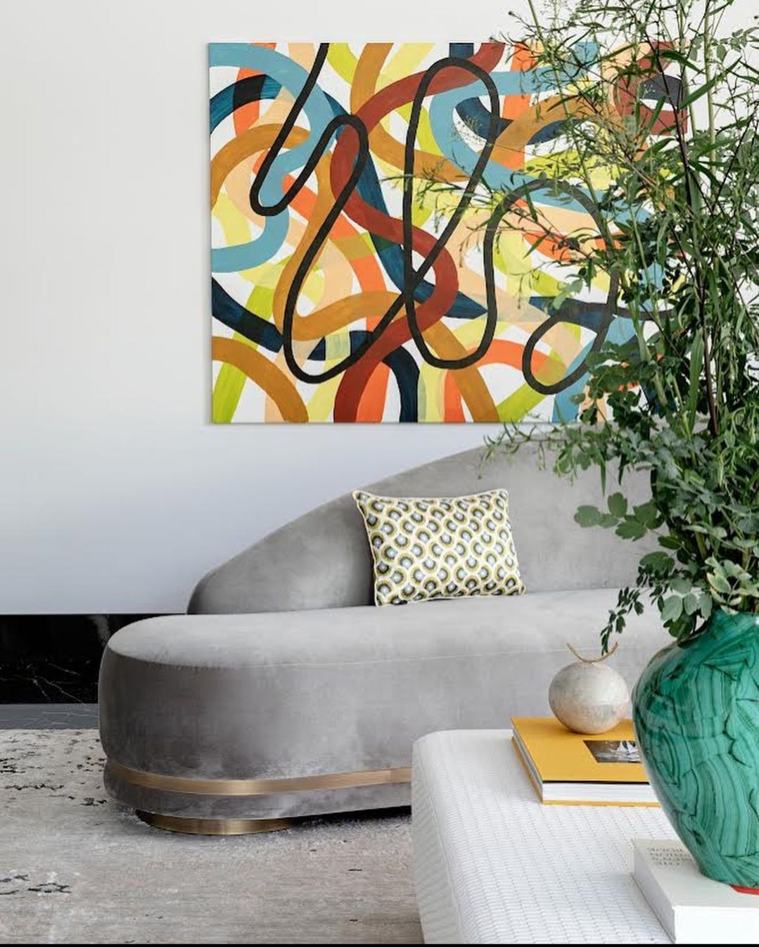 Modern living room with abstract colorful painting, gray curved sofa with patterned pillow, green vase on a white table with books and decorative objects, and greenery.