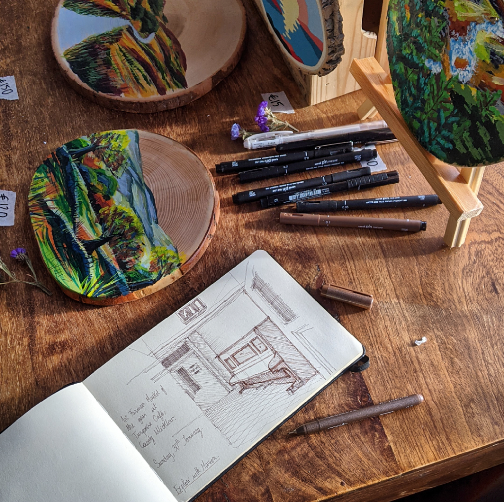 A wooden table displays completed landscape artwork on circular wooden plaques, a sketch of an interior scene in a notebook, and various black and brown pens and markers. There are small flowers and price tags near the art pieces.