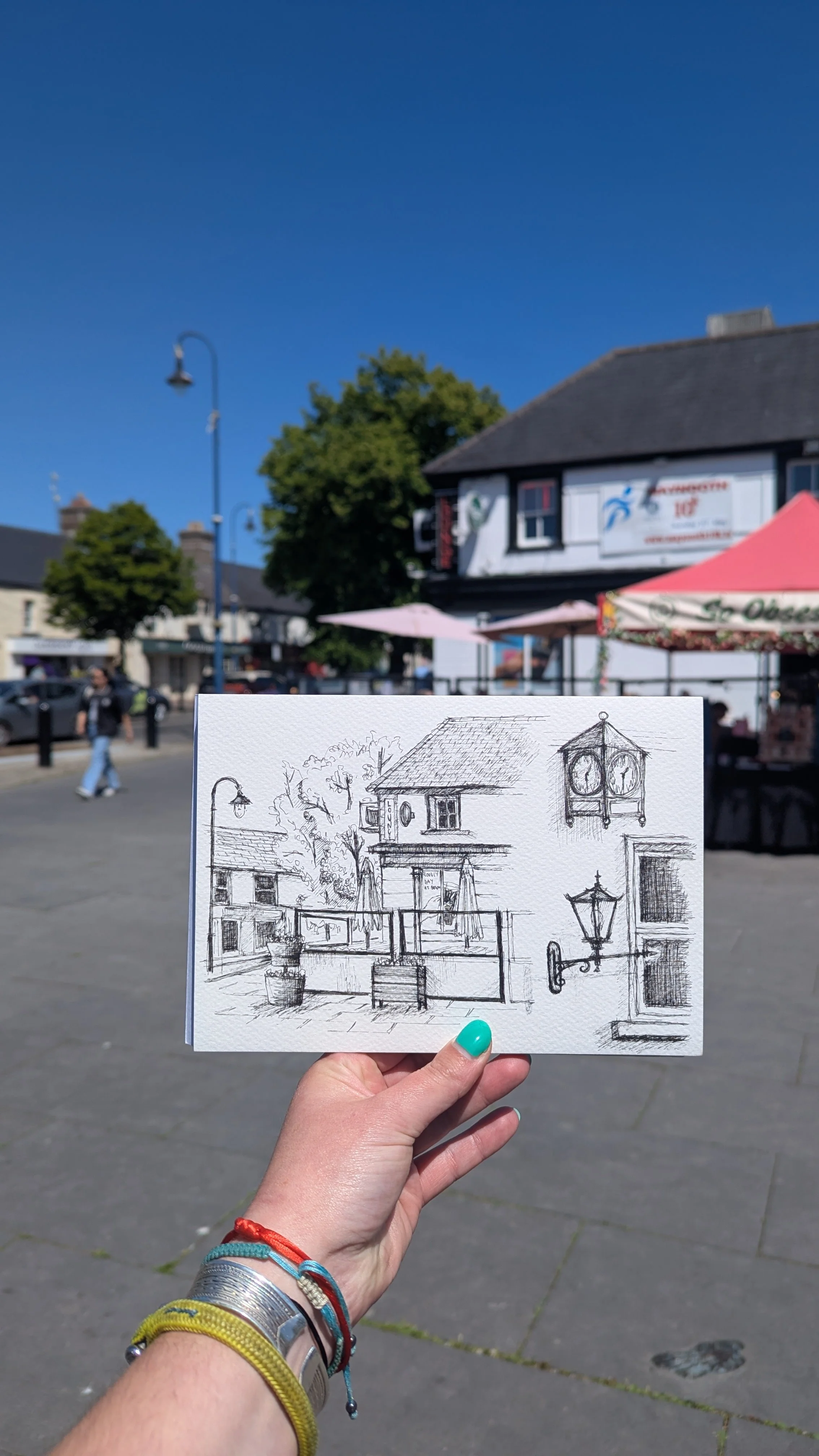 A hand holding a sketch of a town scene with buildings, trees, and street lamps, backdropped by a real town square with shops, a tree, umbrellas, and pedestrians on a sunny day.