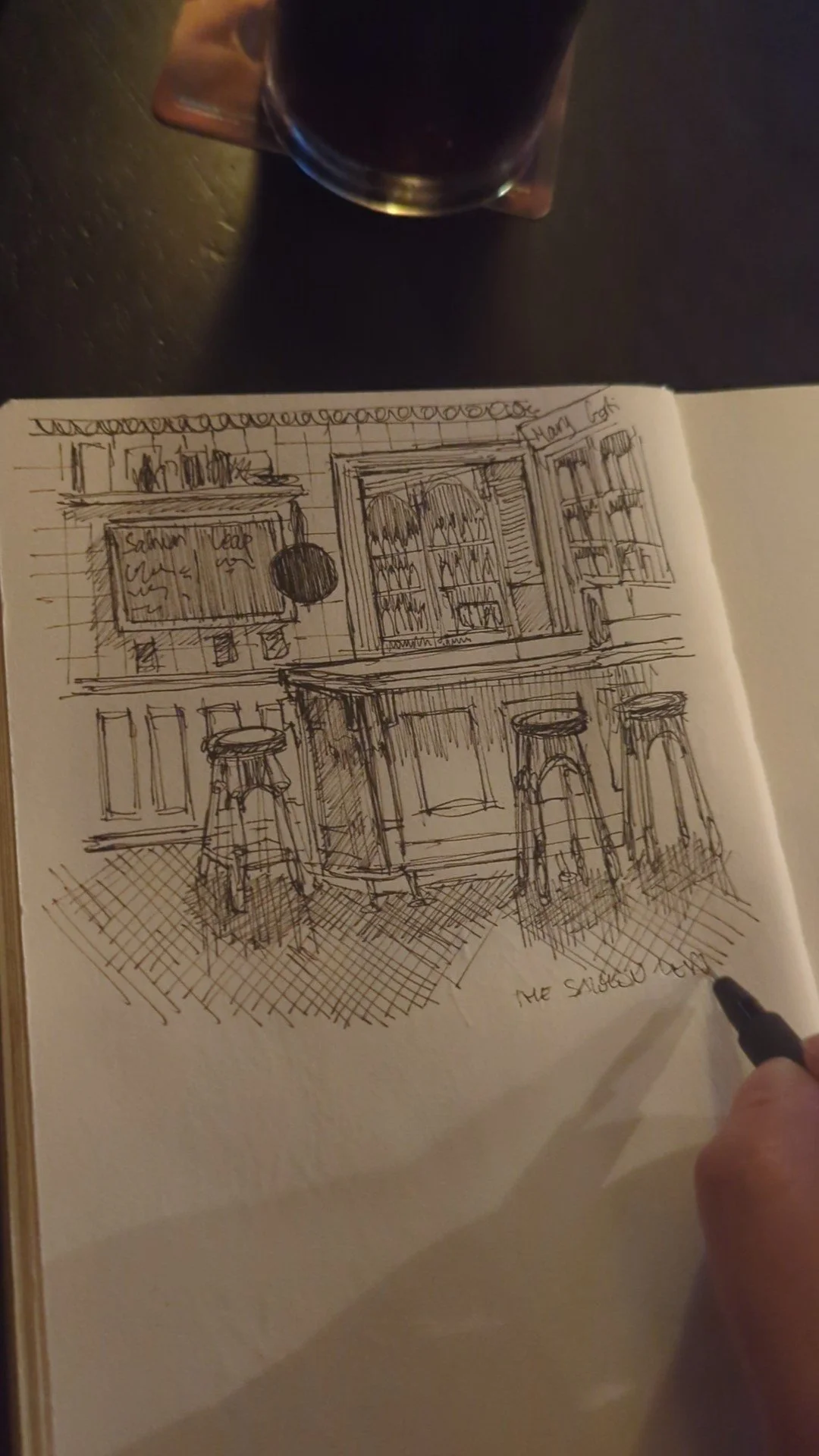 Sketch of a bar interior with shelves of bottles, a flat-screen TV, and three bar stools in front of a counter. A person is holding a pen in the lower right corner.