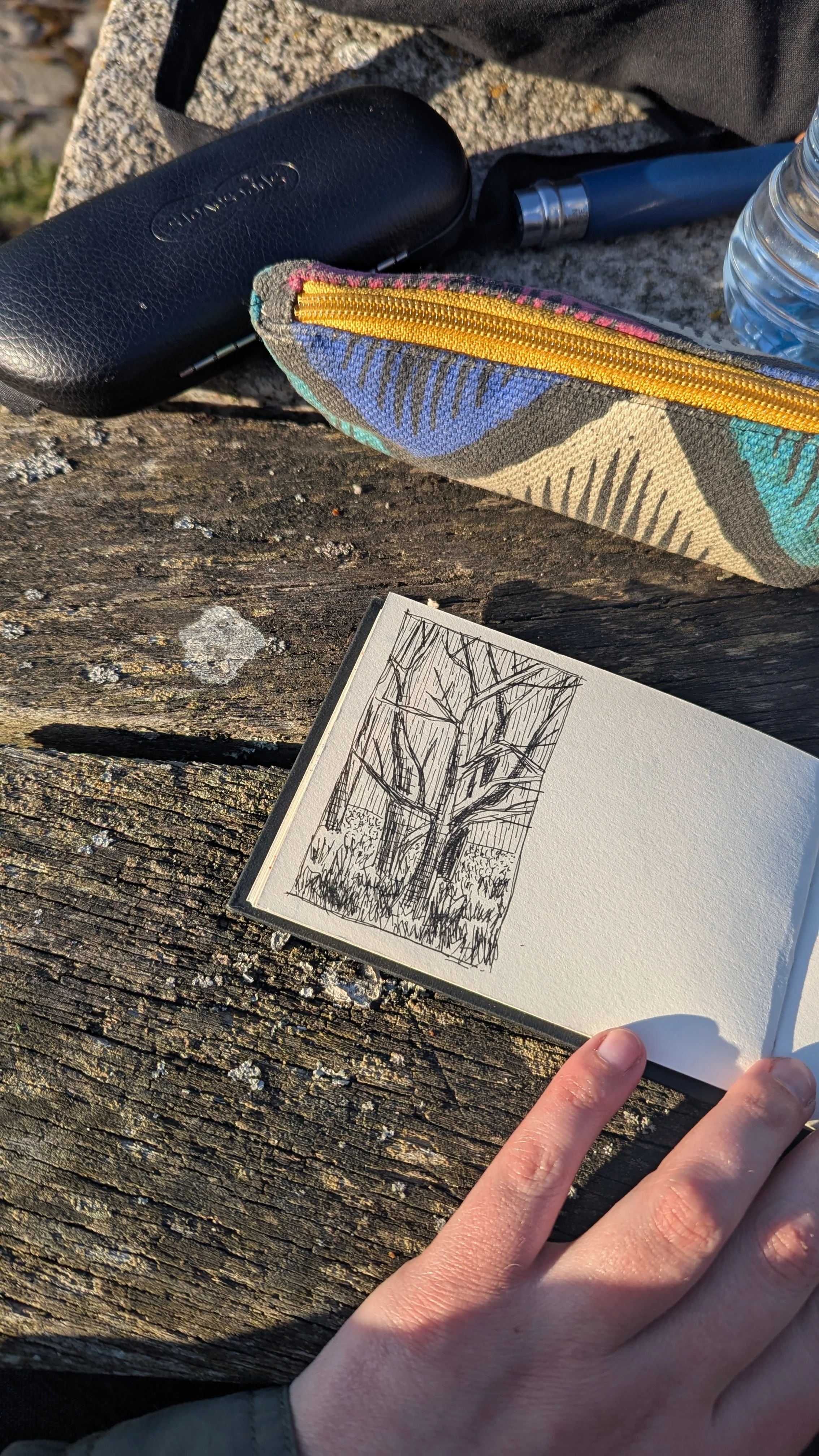 An open sketchbook displaying a black and white drawing of a tree, placed on a weathered wooden surface. A finger is touching the bottom right corner of the sketchbook, with a black textured case, a patterned pouch with yellow zipper, a blue object,