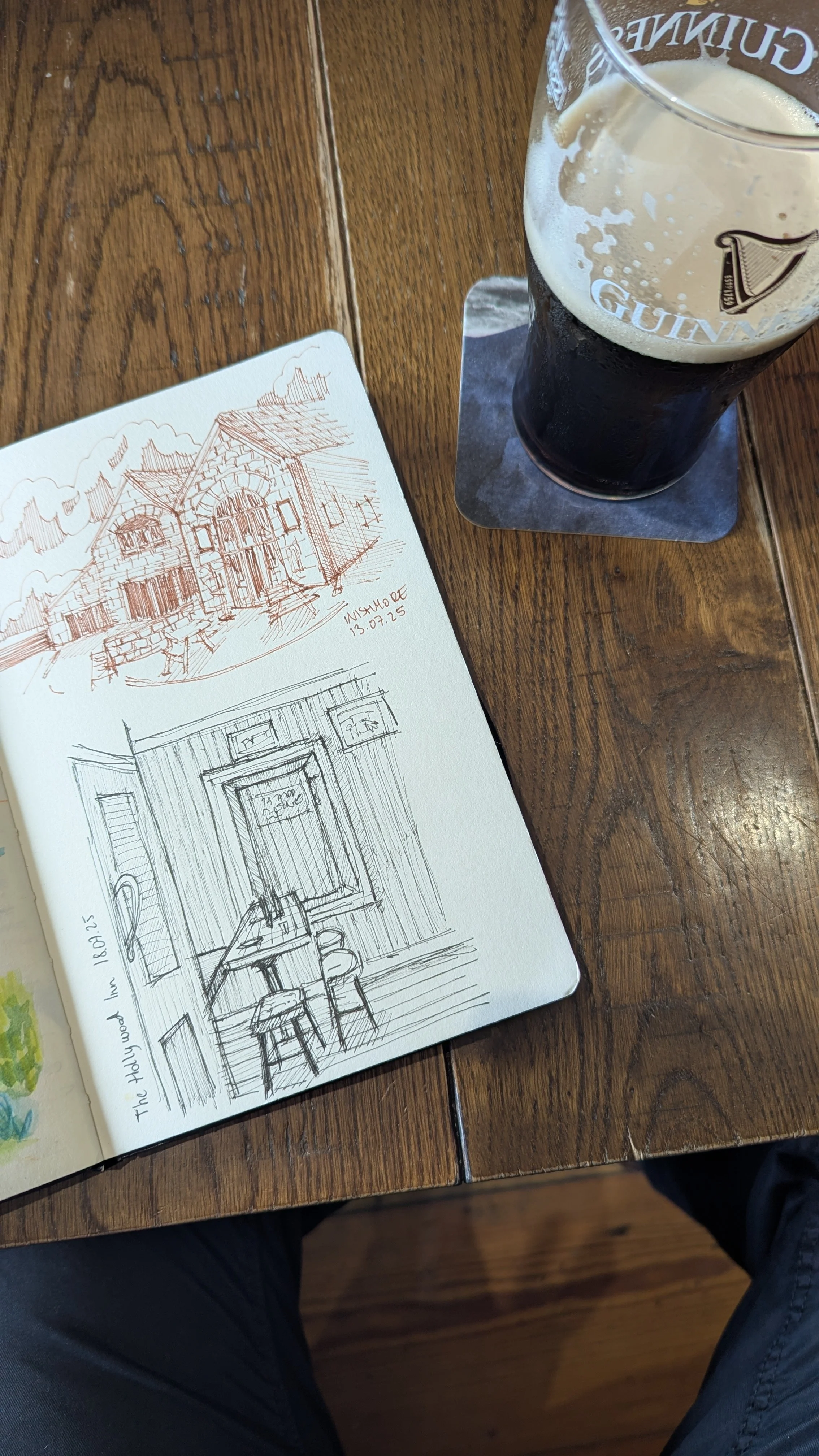 A sketchbook with two sketches of a house and an interior room, a pint of Guinness beer on a coaster on a wooden table, and part of a person's legs in black pants.