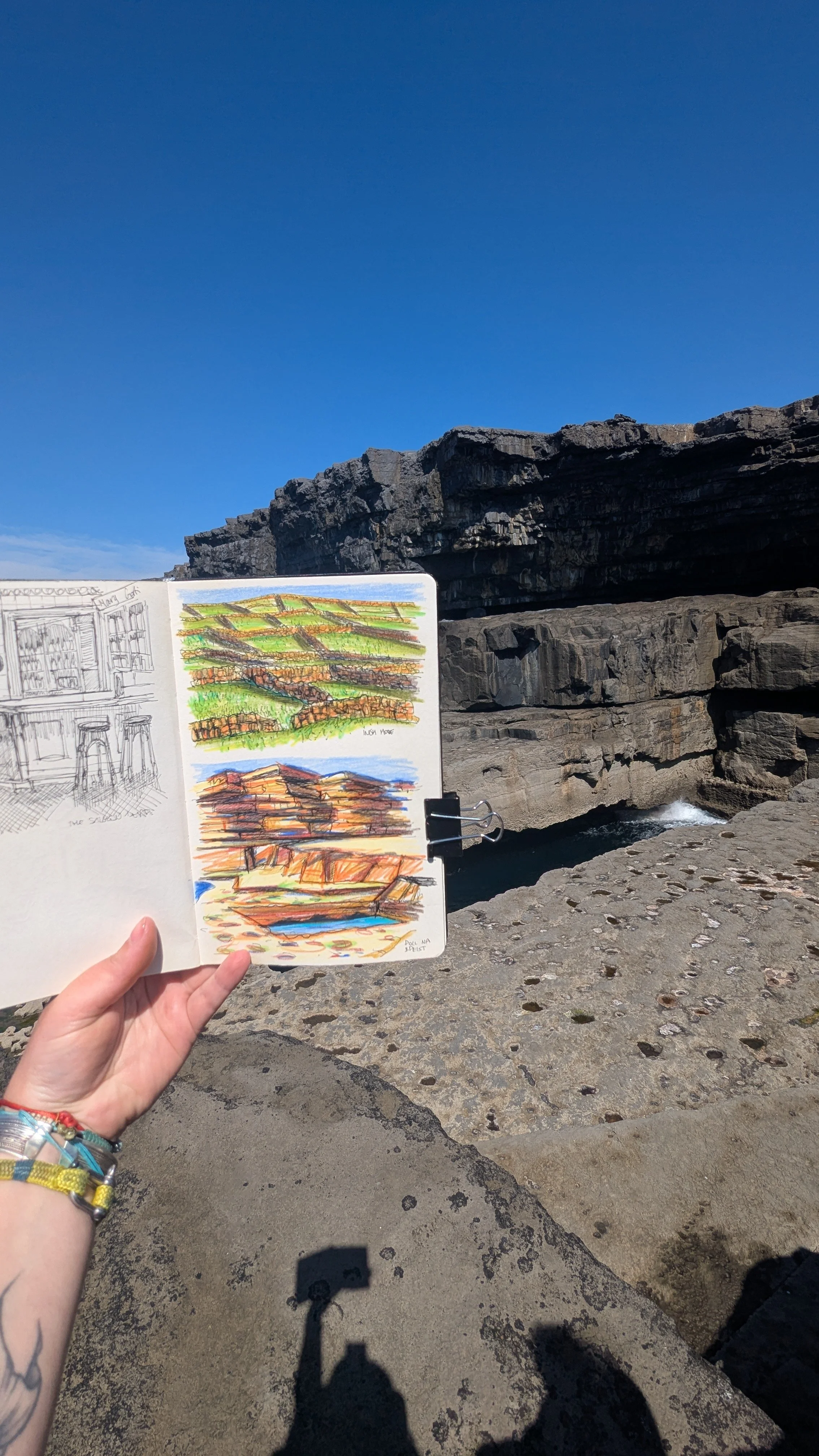 A person holding an open sketchbook with landscape sketches, standing on a stone surface near rugged coastal rocks under a clear blue sky.