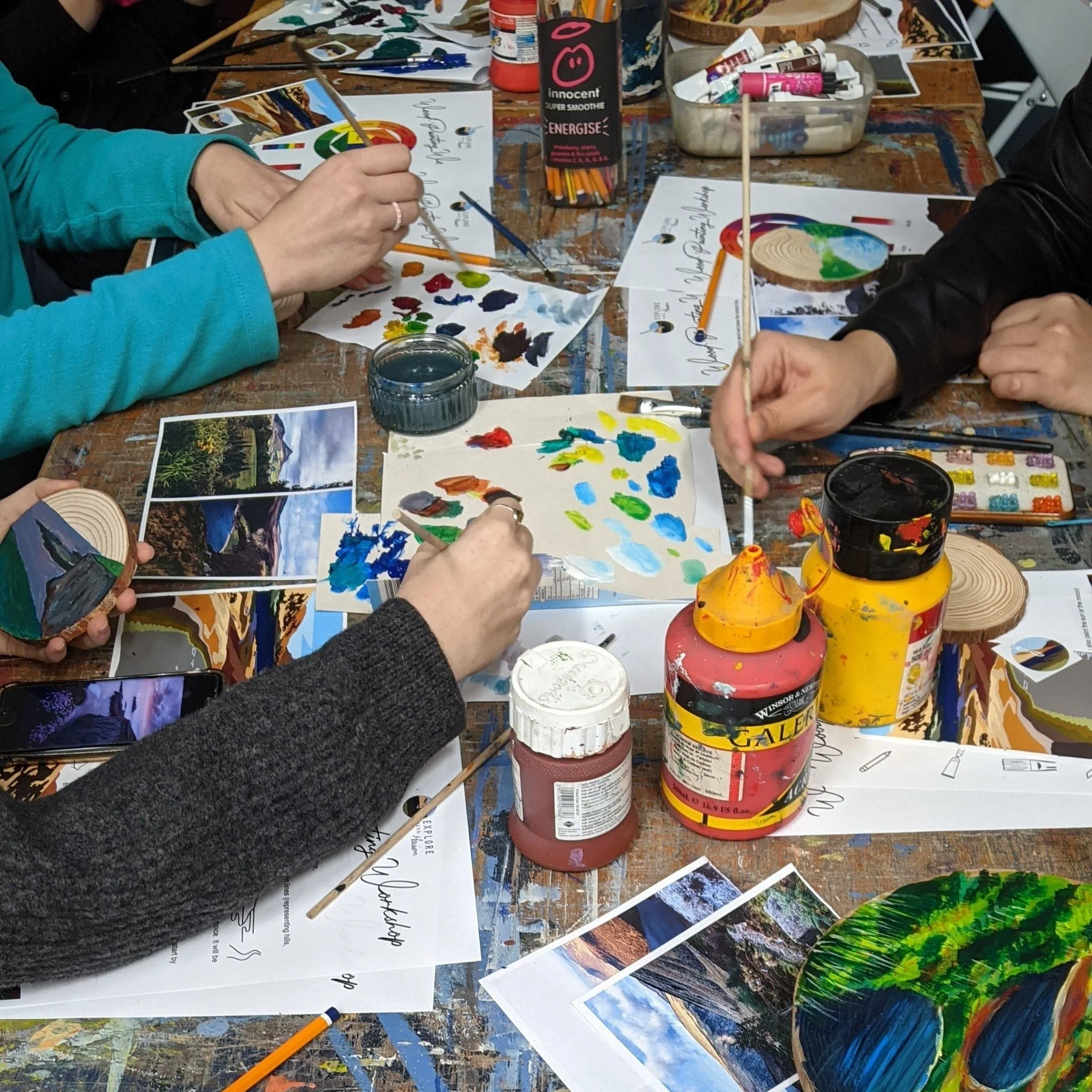 People painting wooden crafts at a table, surrounded by paints, paintbrushes, and landscape photos.