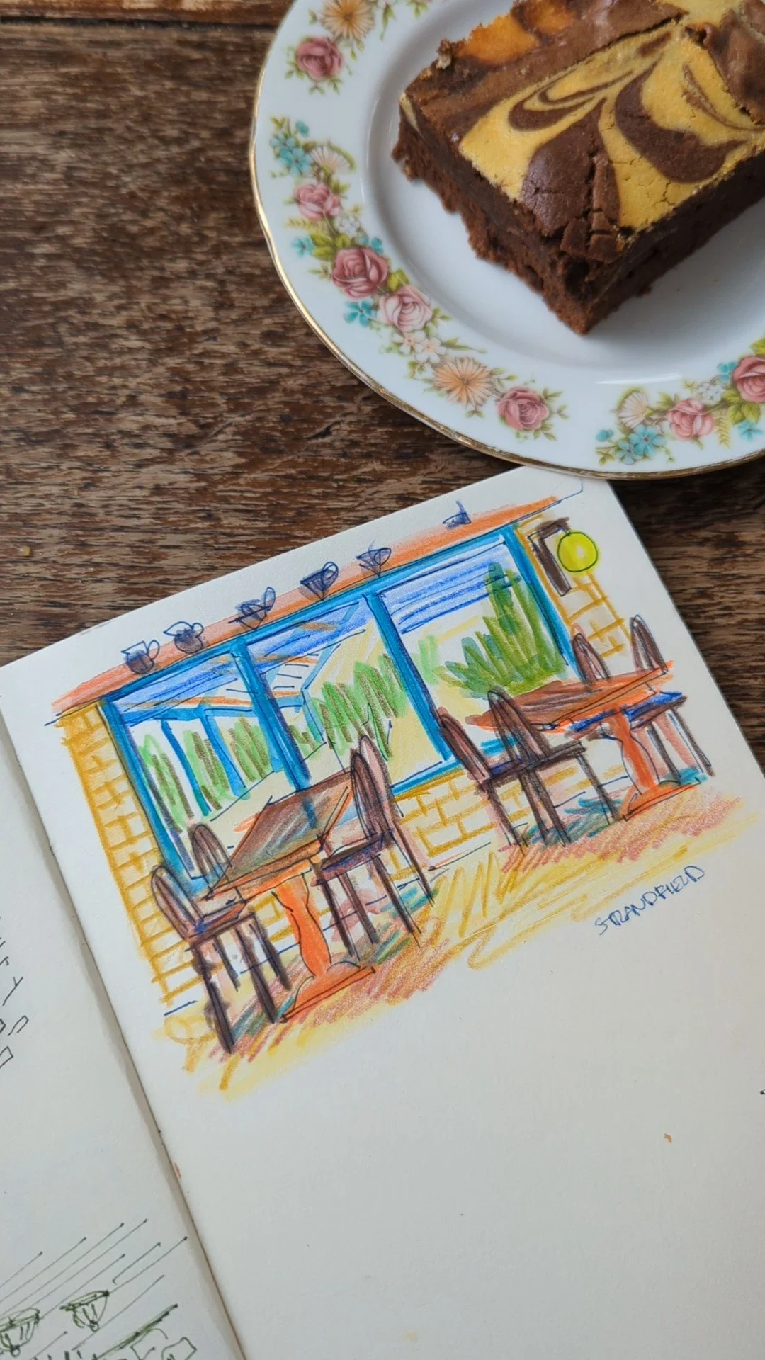 A decorated marble brownie on a floral china plate, next to a colorful sketch of a patio with tables, chairs, and greenery.
