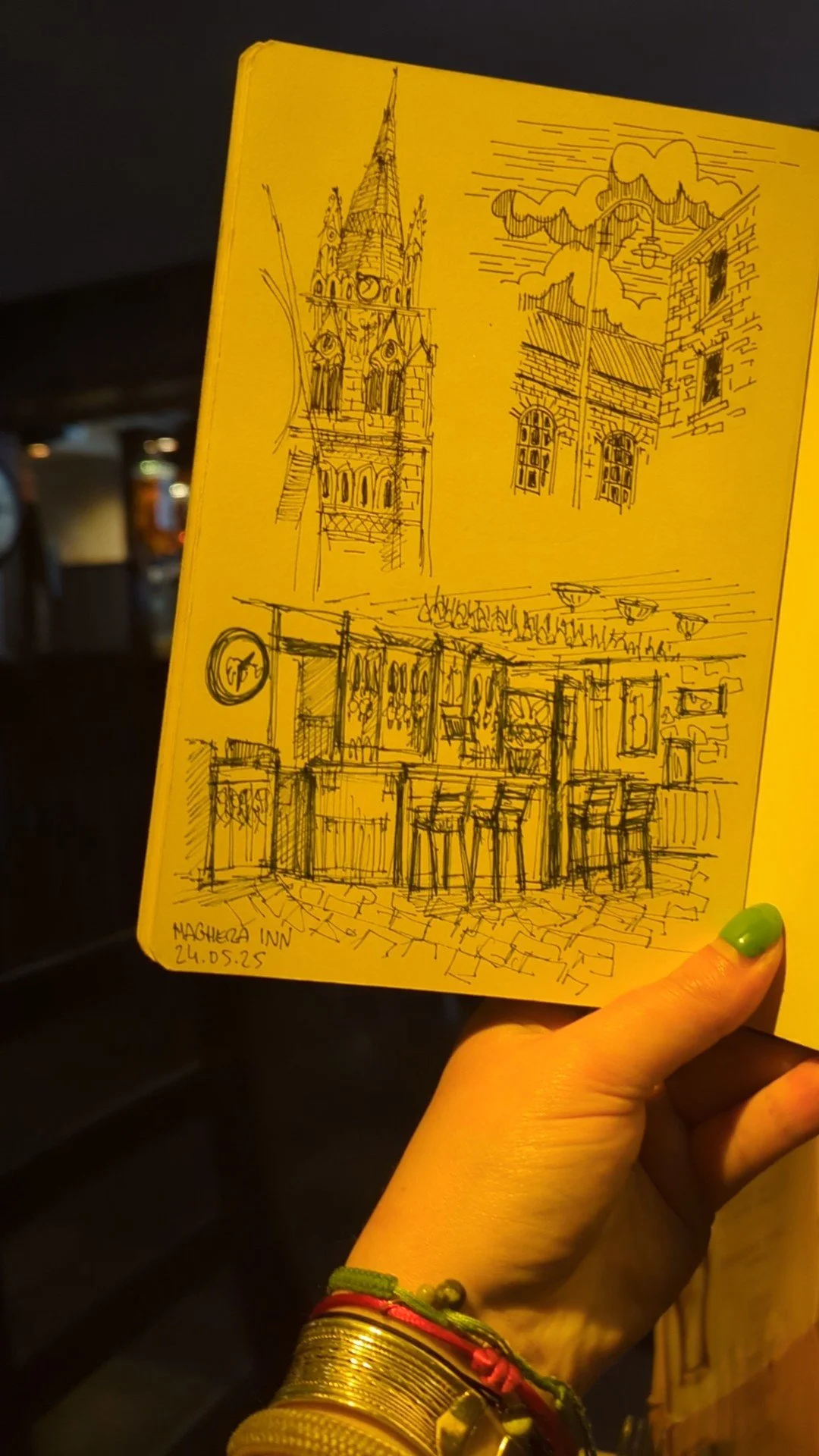 A yellow notebook with sketches of a clock tower, a building with windows, and a bar interior with high chairs and hanging lights, held by a person with gold jewelry and a green nail.