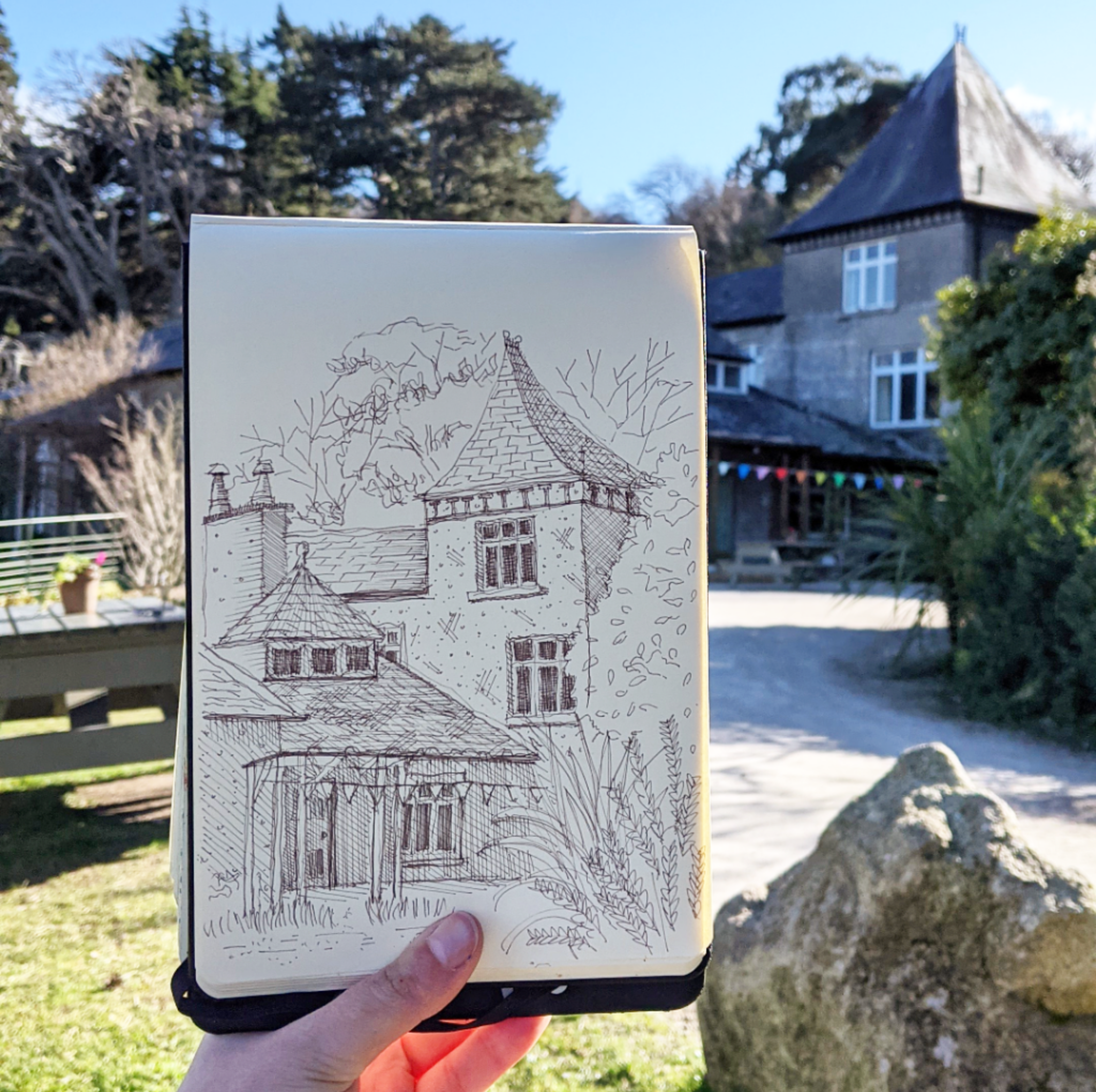 A person holding a sketchbook with a drawing of a house and trees, with a historic house and garden visible in the background on a sunny day.