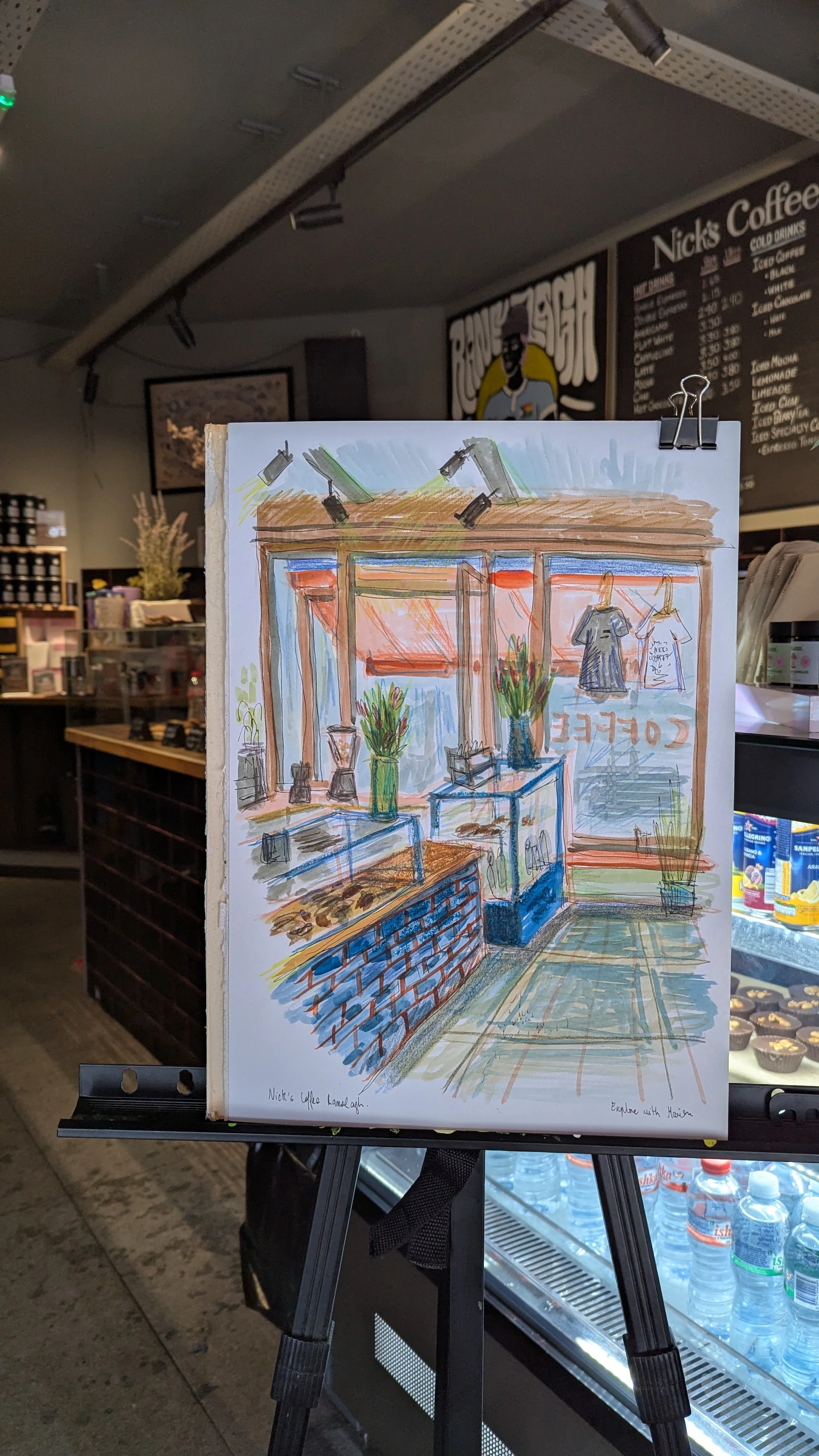 A colorful hand-drawn sketch of the interior of Nick's Coffee. The sketch shows a counter with vases and plants, a display case, and hanging shirts in front of large windows with a view outside. The real coffee shop interior is visible in the backgro