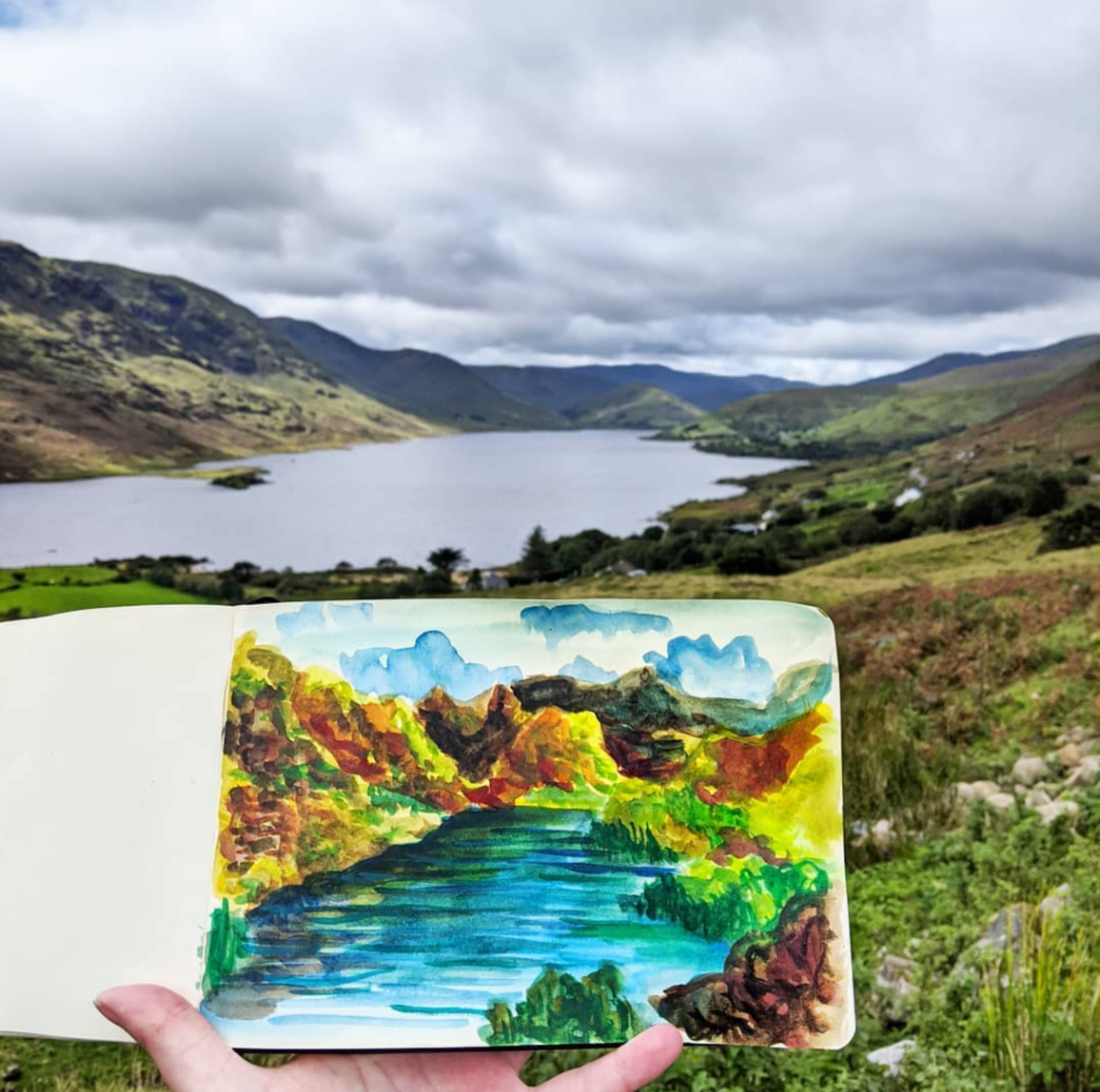 A lake surrounded by green hills under a cloudy sky, with a hand holding an open watercolor sketchbook displaying a colorful landscape painting of the same scene.