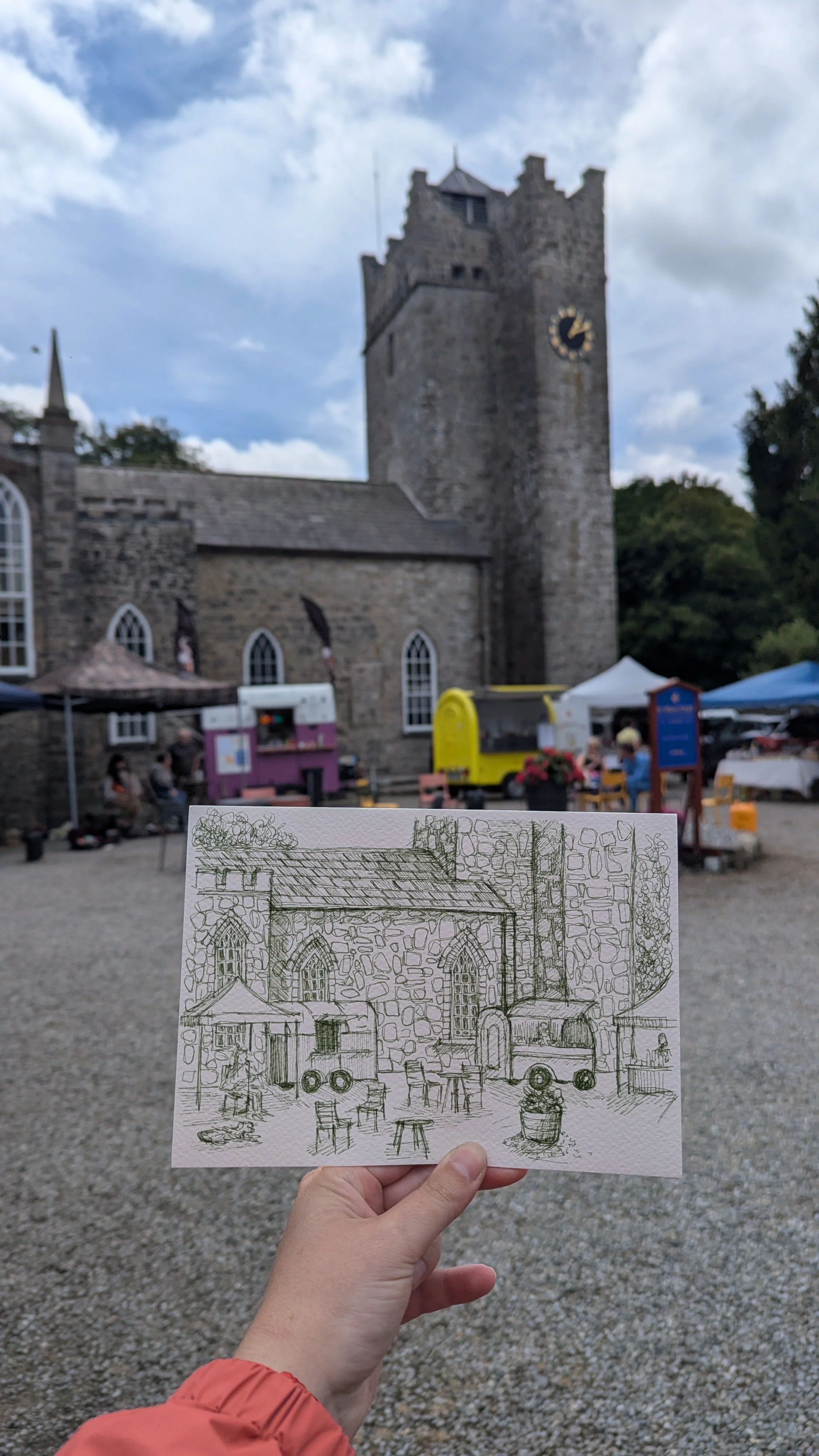 Person holding a sketch of a stone church and outdoor seating in front of the actual stone church with a clock tower.