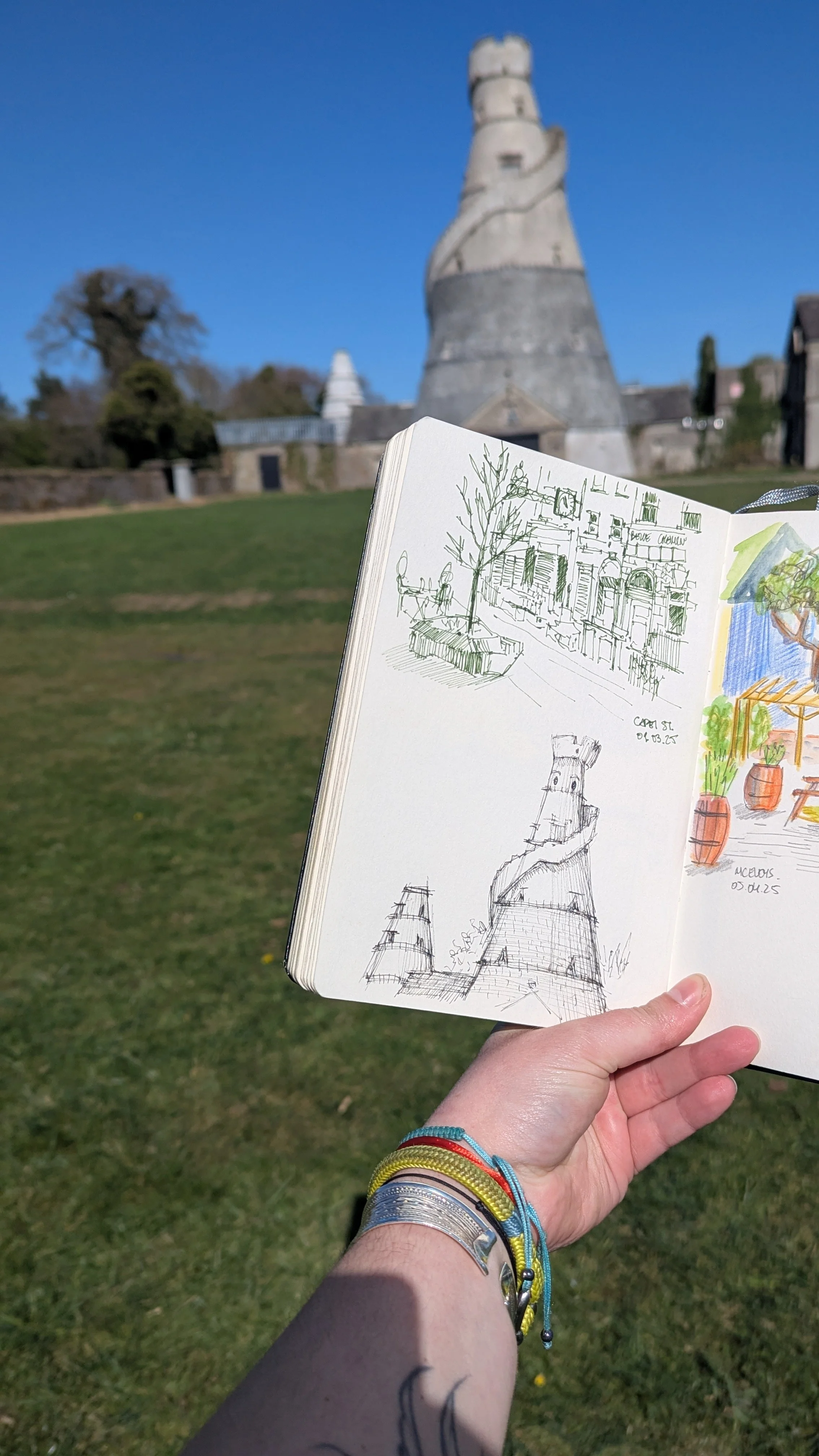 A person holding a sketchbook with drawings of a tower and street scene in front of a historical tower under a clear blue sky.
