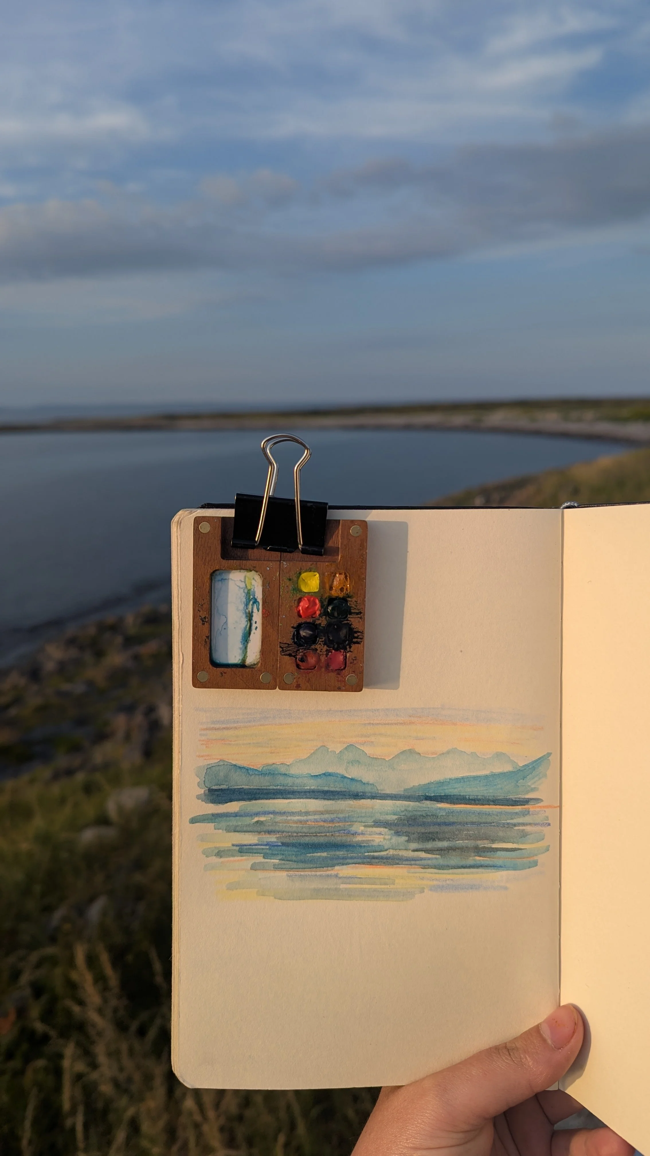 A person holding an open sketchbook with a watercolor landscape of mountains and water, set outdoors near a body of water with scenic sky in the background. There is a small palette and a paintbrush attached to the top of the sketchbook using a binde