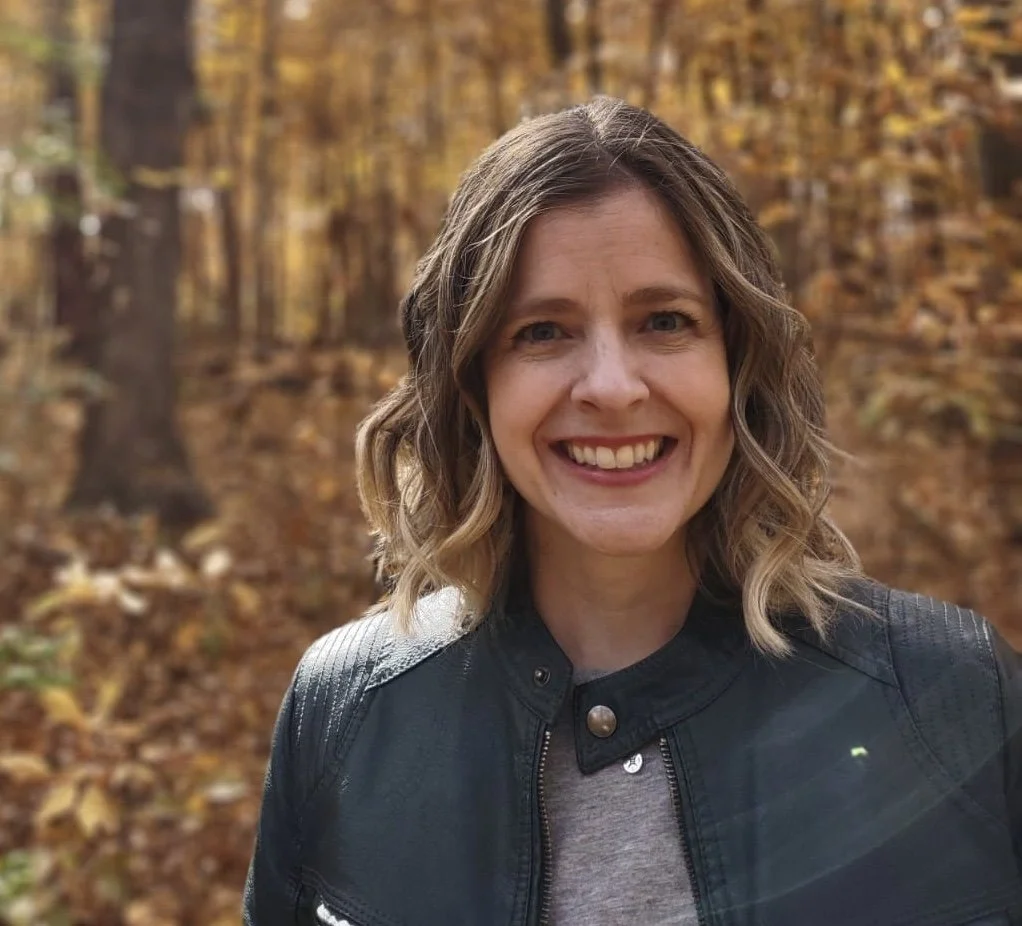 A woman smiling outdoors in a forest with autumn-colored leaves, wearing a black leather jacket and a gray top.