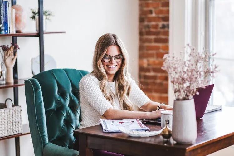 A woman with long blonde hair and glasses smiling while working on a laptop at a wooden desk in a well-lit room with brick wall and shelves with decorative items giving a human design reading.