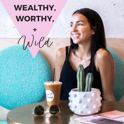 A woman sitting at a table in a cafe with a cactus plant, a cold drink with a straw, and a pair of sunglasses, smiling and looking away. The background includes pink and white textured wall art with the words 'Wealthy, Worthy, + Wild'.
