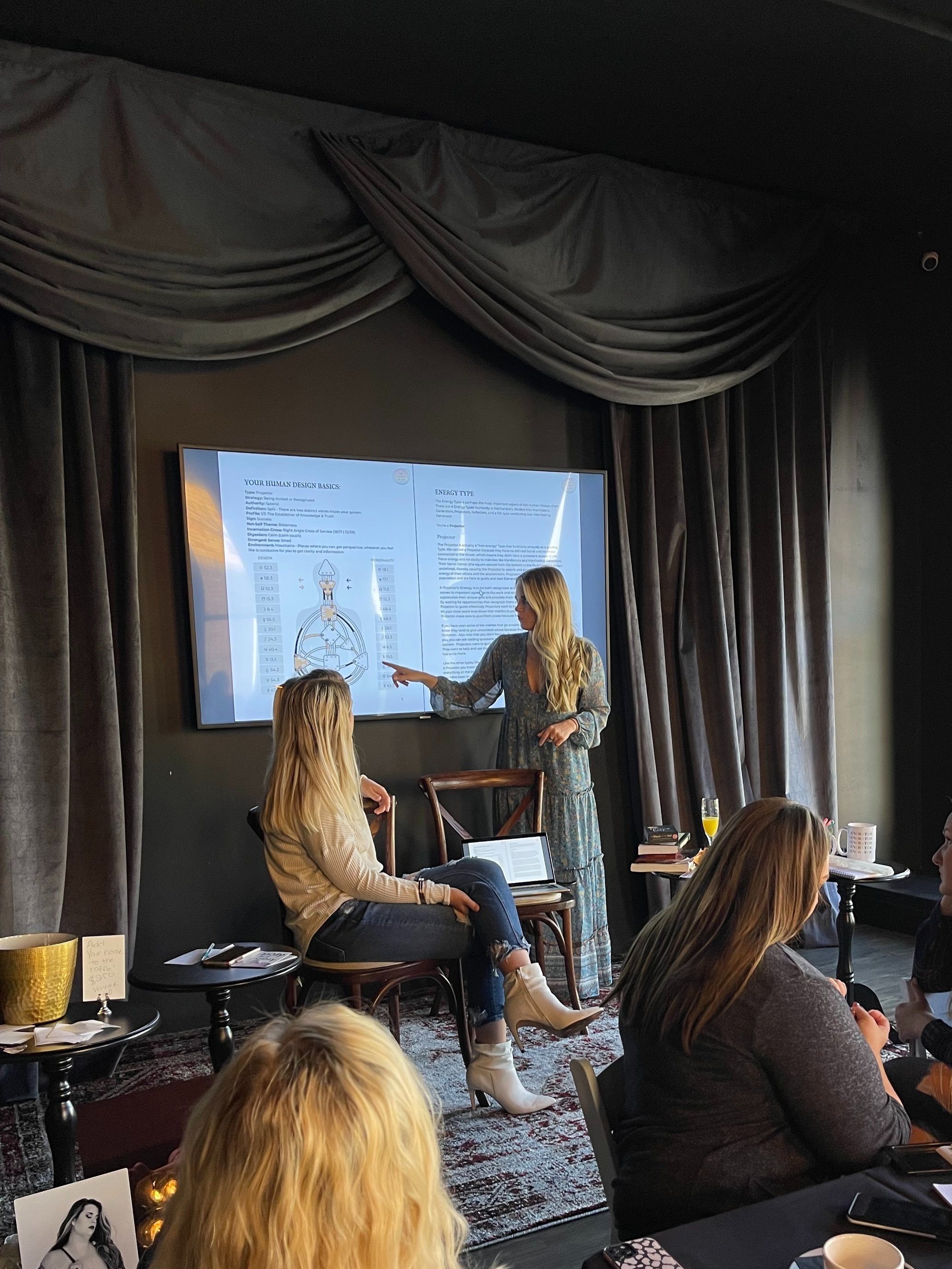 A woman giving a presentation to a group of women seated at a table in a darkened room with large curtains. The presenter is pointing at a large screen displaying diagrams and text.