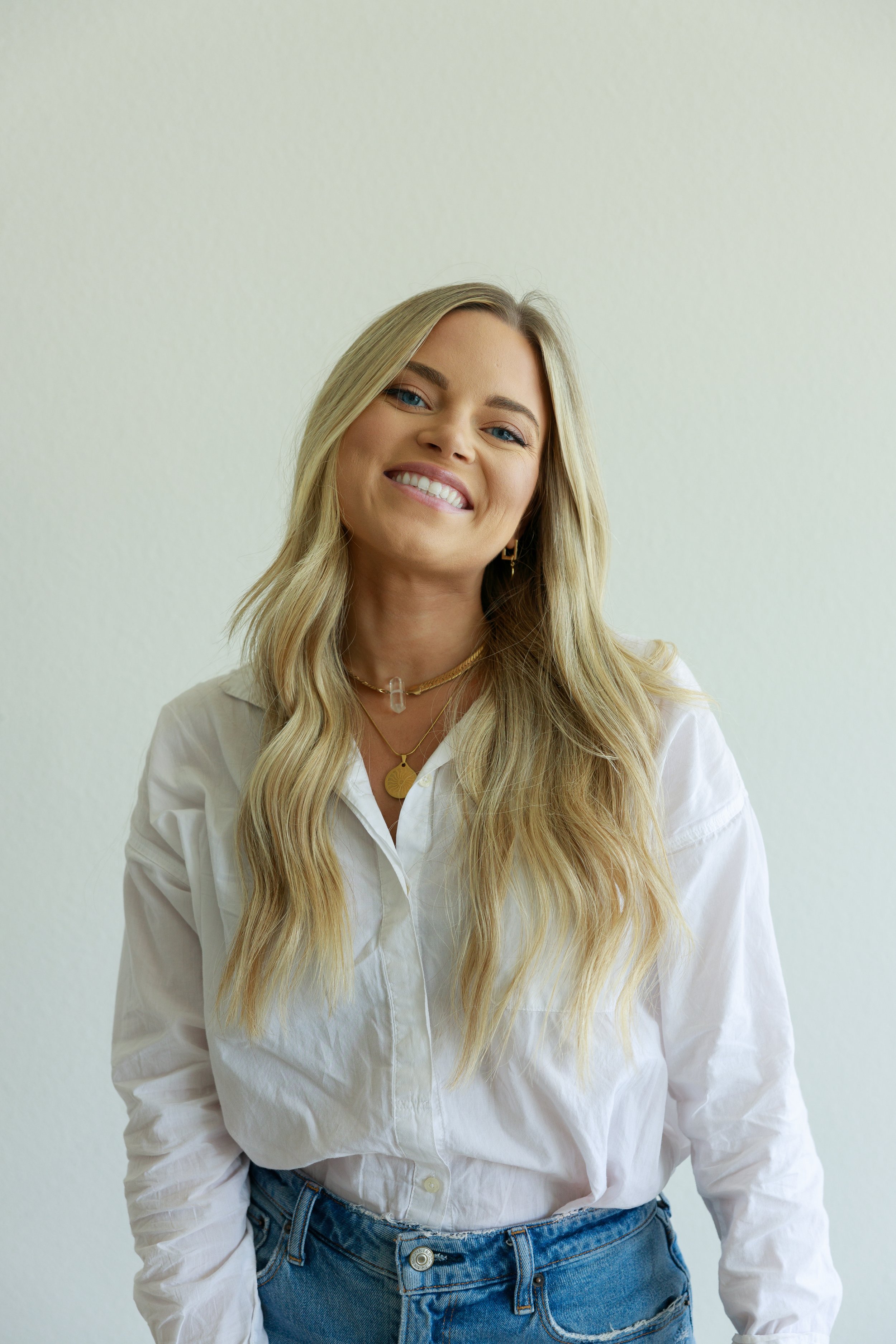 A young woman with long blonde hair smiling, wearing a white button-up shirt and blue jeans, standing against a plain light-colored background.