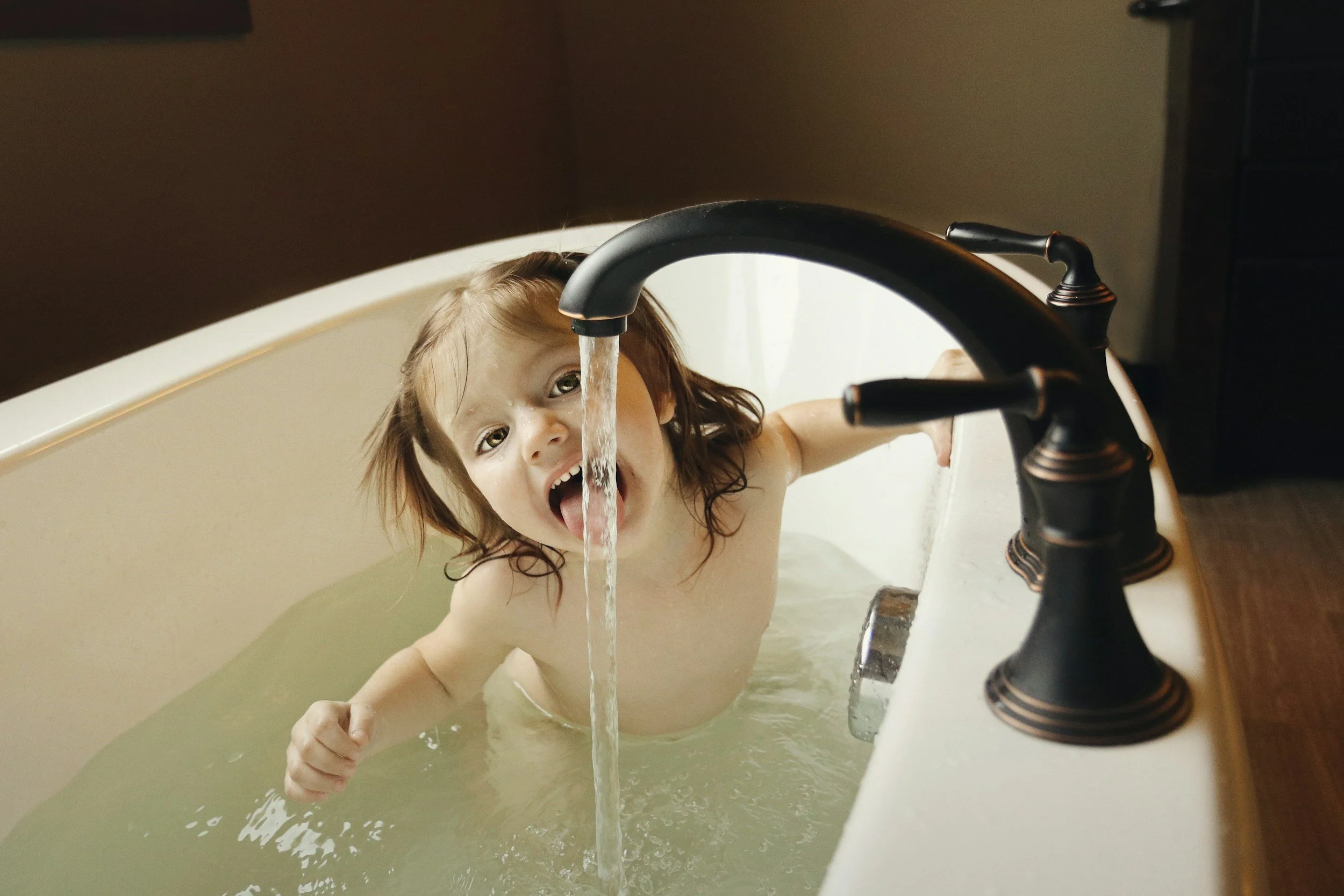 A young child with wet hair is sitting in a bathtub filled with water, playing under a running faucet. The child has an excited expression and is sticking out their tongue.