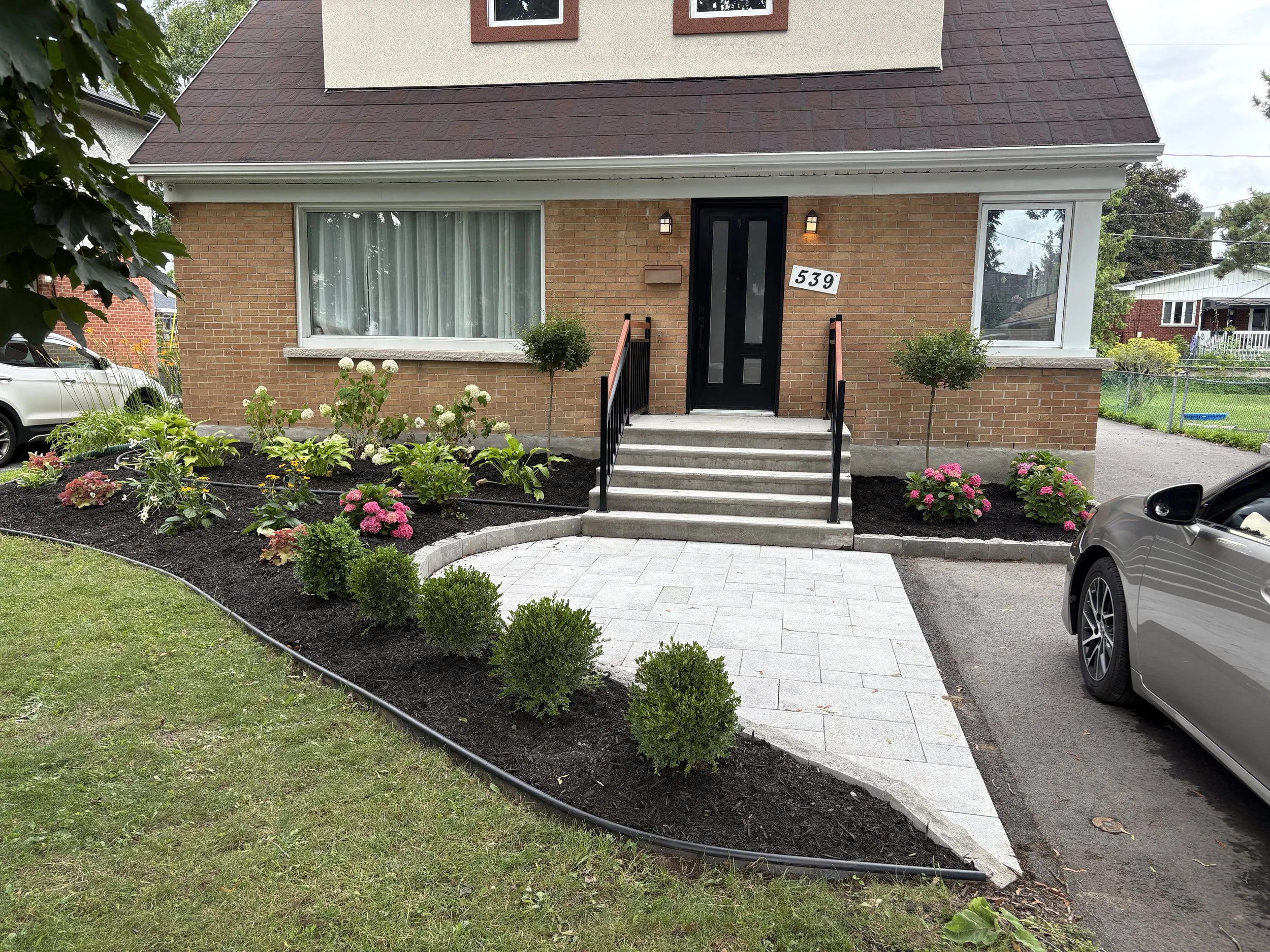 Front yard of a brick house in Ottawa with elegant landscaping. A garden with bushes and flowering plants adorns a curved hardscaped walkway leading to the entrance.