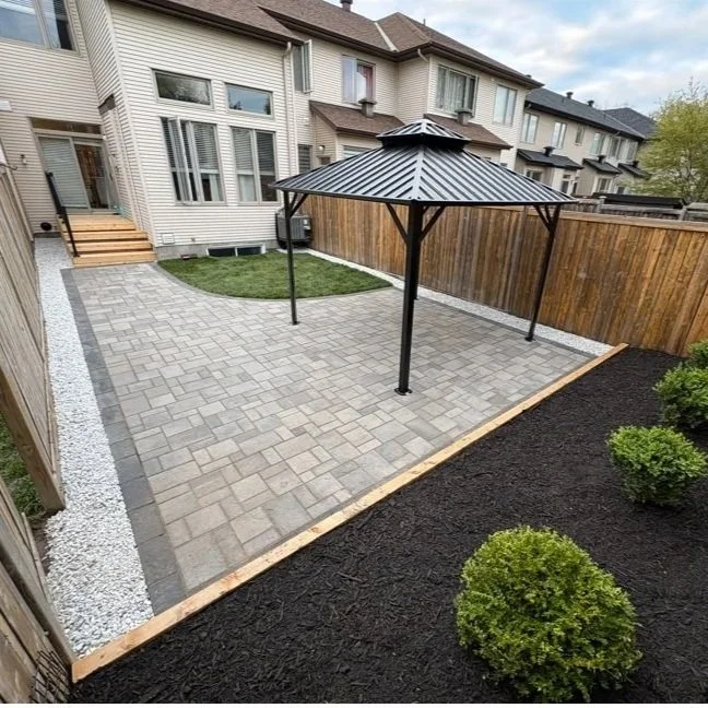 Backyard with a curved paver patio, metal gazebo, small patch of grass, and flower bed with boxwood shrubs and mulch.