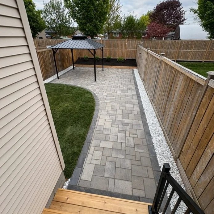 A backyard patio with a curved paver walkway, a wooden fence, a small grass patch, and a metal gazebo.