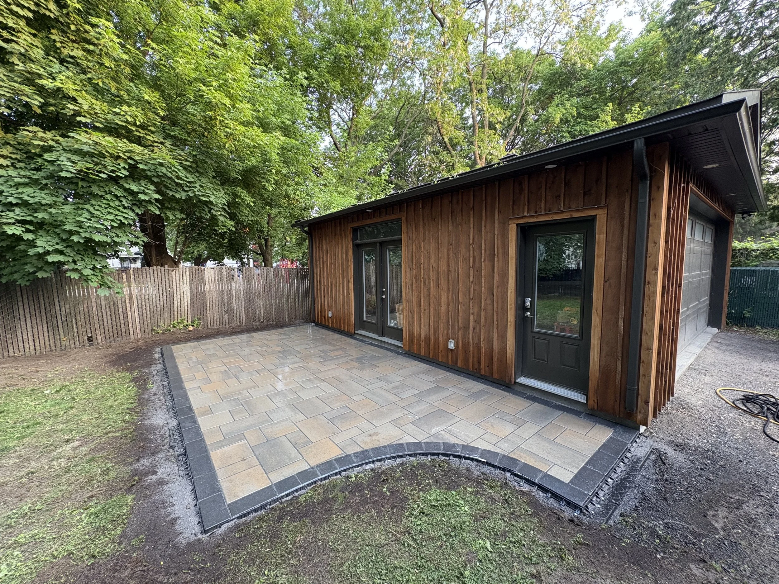 A small wooden backyard shed adjoining a paver patio with an elegant design and a sharp black border, surrounded by a garden with trees and a wooden fence.