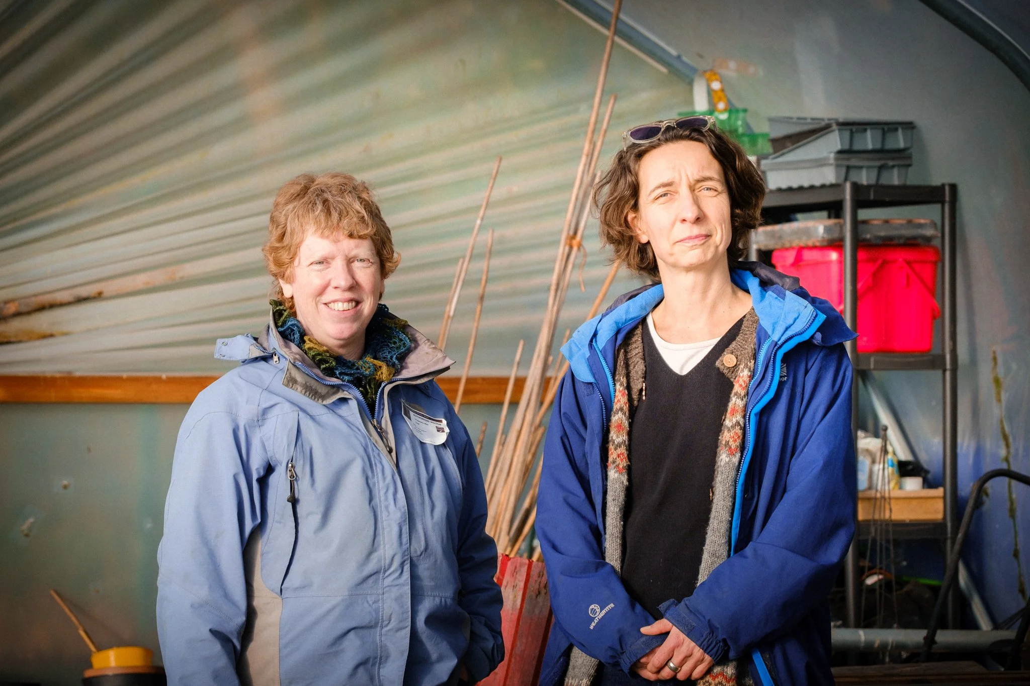 Two women wearing blue jackets inside a shed or garage, standing in front of gardening tools and storage shelves.