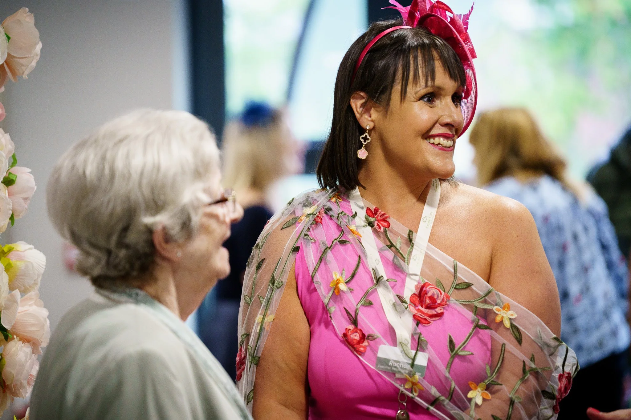 A woman in a pink dress and a pink fascinator headband is smiling, standing next to an elderly woman in white, at an indoor event in Chester