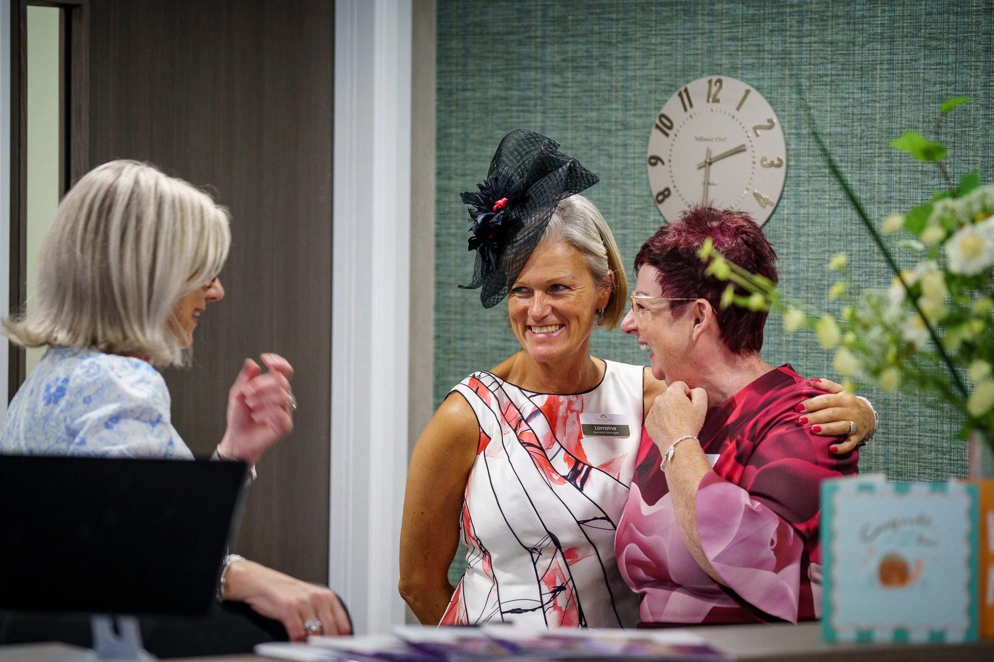 Three women interacting in an indoor setting; one woman wearing a black fascinator hat, one with glasses and short hair, and another with red hair and a red patterned dress; a clock on the wall shows 2:15; a flower arrangement partially visible in th