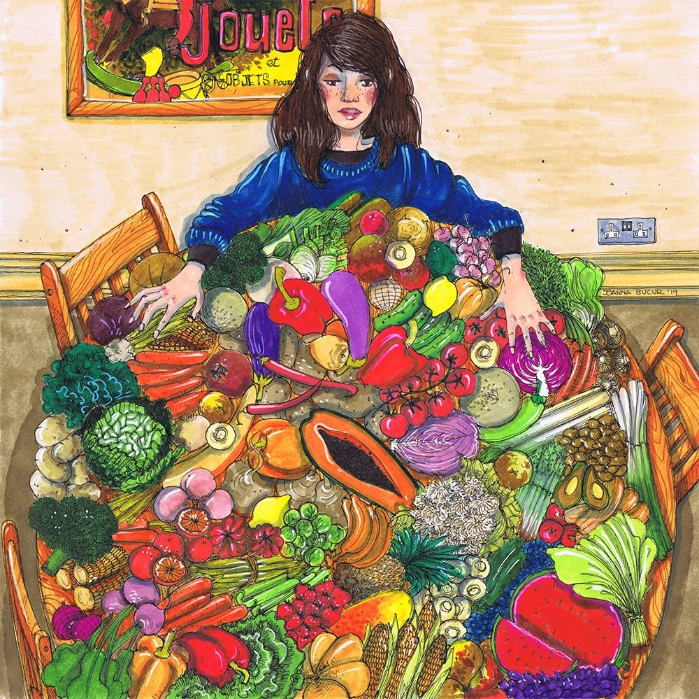 A woman with short brown hair and a blue sweater sitting at a table overflowing with colorful vegetables.