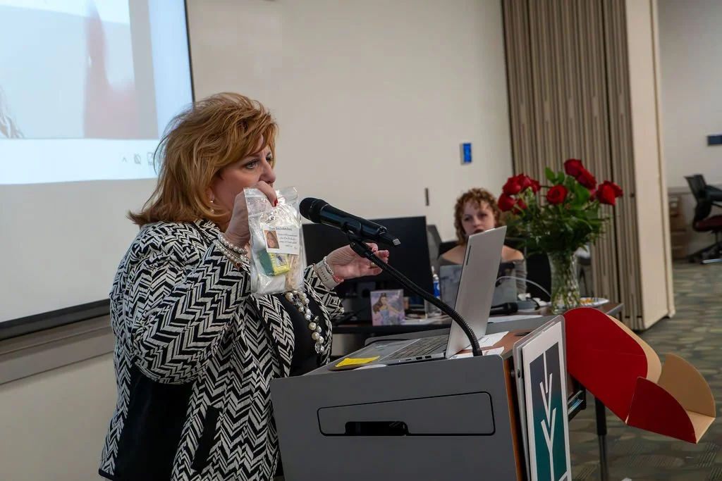 A woman standing at a podium holding a plastic bag in a conference room, while another woman sits behind her with a laptop and a large bouquet of red roses.