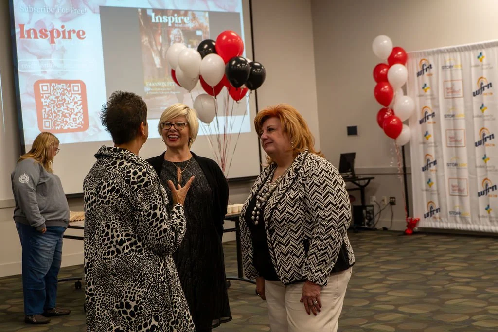 Three women are engaged in conversation in a conference room decorated with black, white, and red balloons, with a large screen and a branded backdrop in the background.