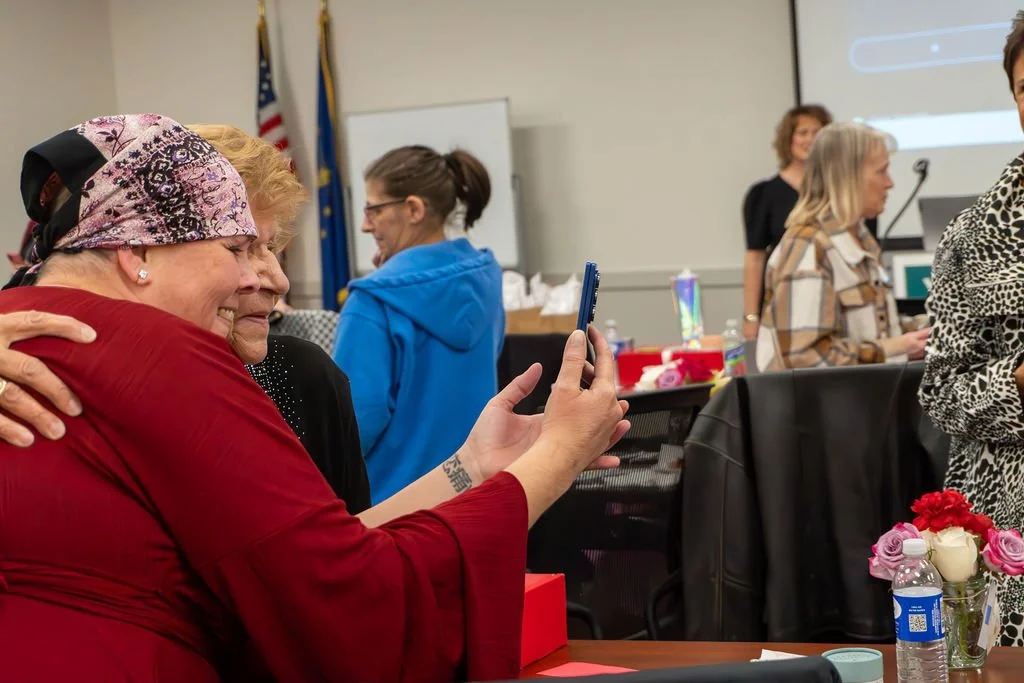 A group of women in a meeting room, one woman in a red shirt and pink headscarf taking a selfie with a woman in black, others engaging in conversations, laptops, flowers, and American flags in the background.