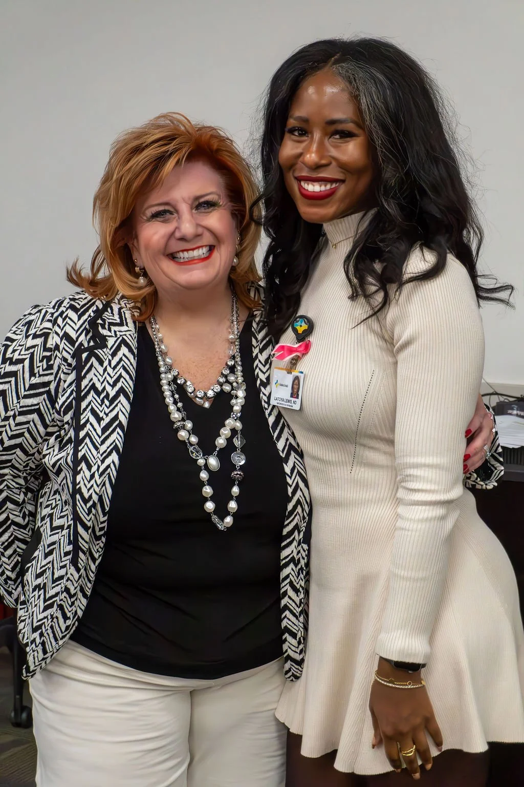 Two women smiling and standing close together, one wearing a black top with a patterned blazer and pearl necklaces, the other in a white dress with employee badge and pink ribbon pin.