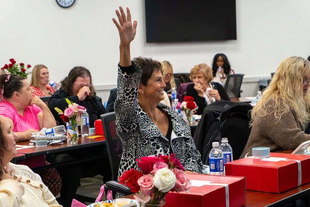 A woman with short brown hair raising her hand during a meeting in a conference room decorated with flowers and water bottles.