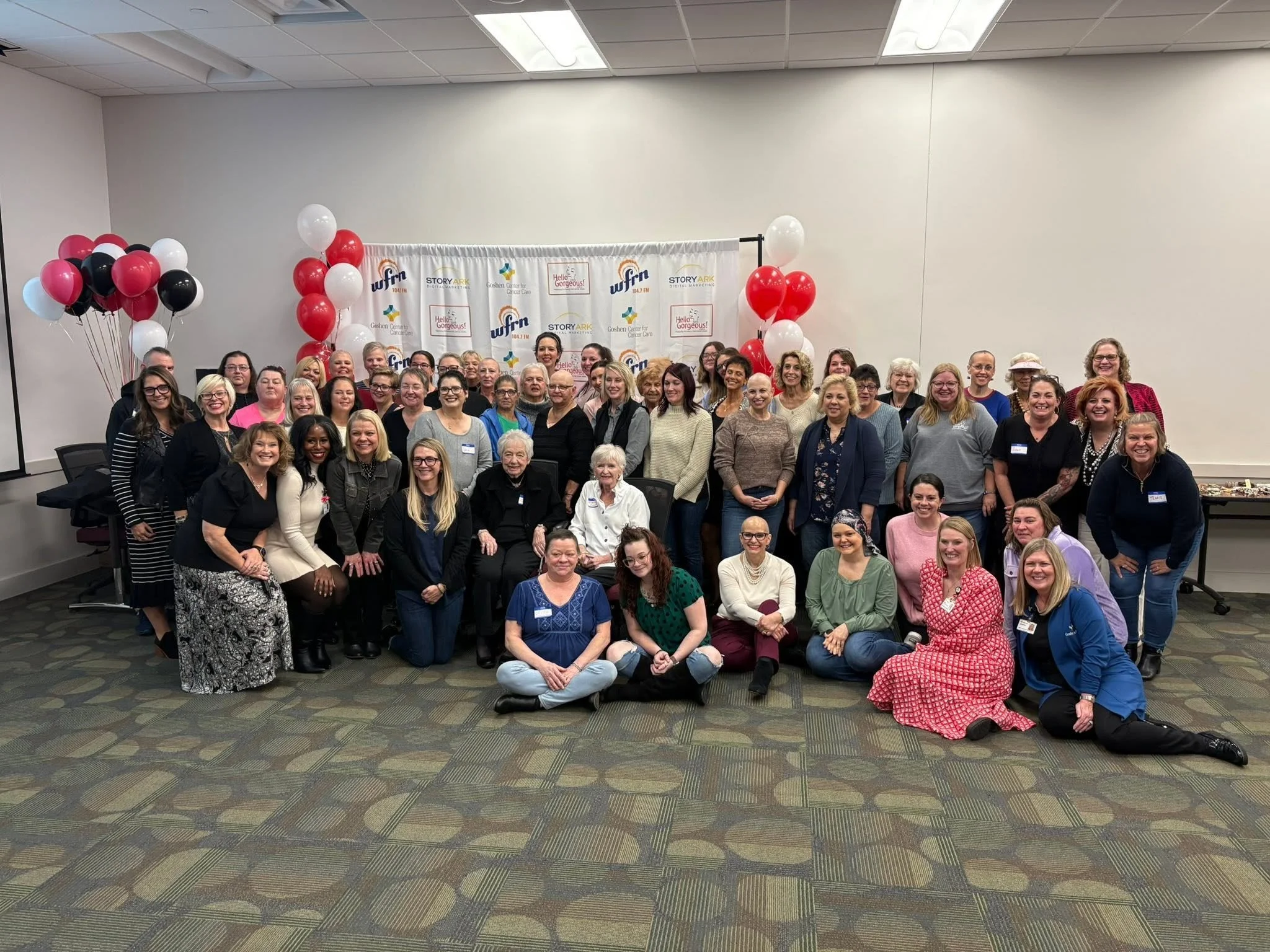 A large group of smiling women and men of various ages gathered in a room for a group photo, with balloons and a backdrop displaying logos and text related to a community or organizational event.