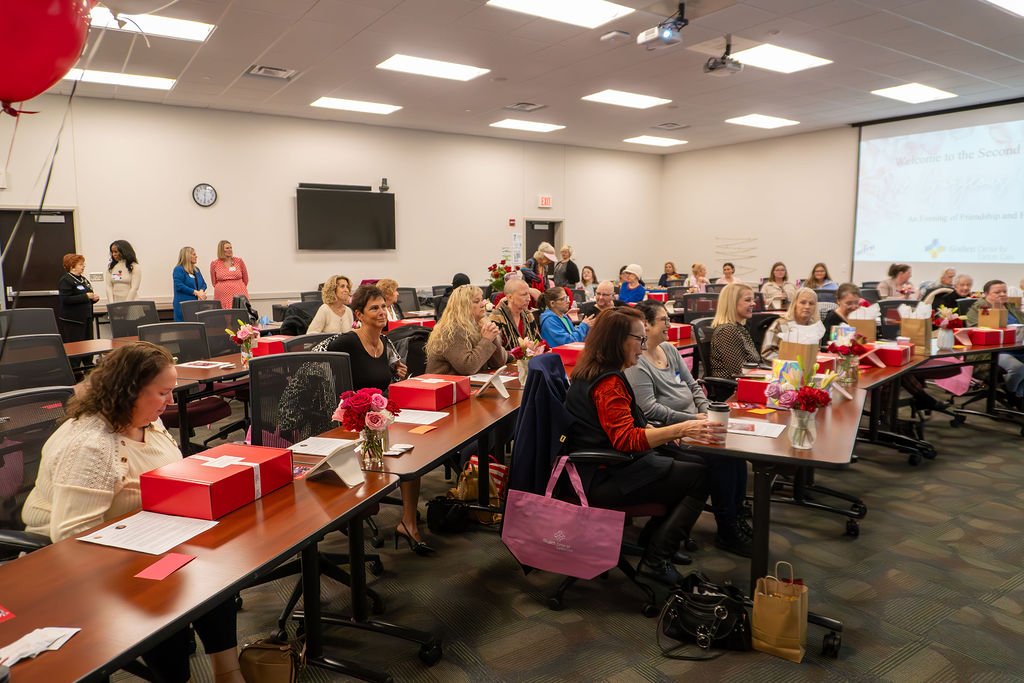 A conference room filled with women sitting at tables decorated with flowers and red gift boxes, attending a presentation on a large screen.